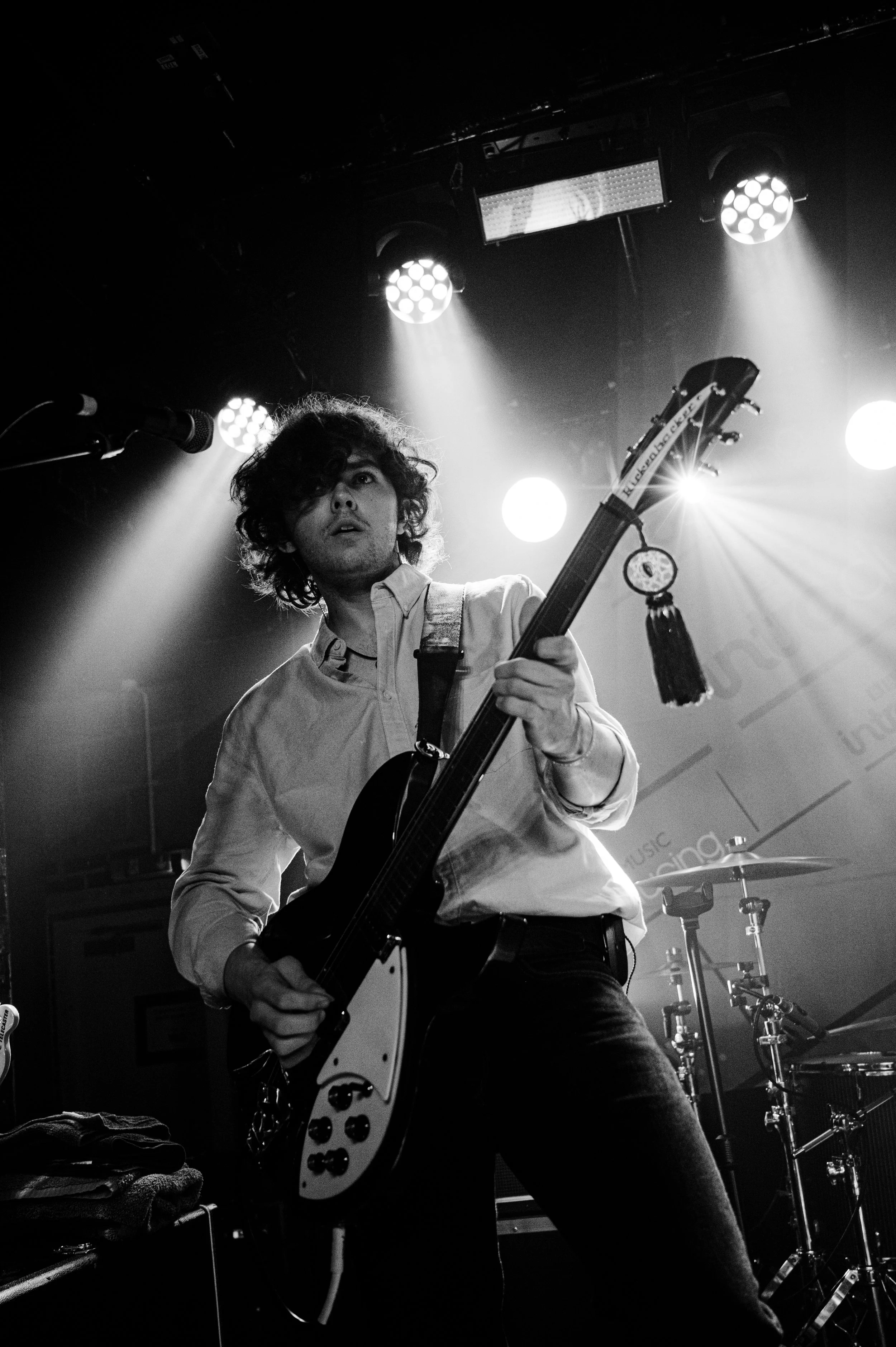 A black and white photo of a young man playing an electric guitar on stage, with stage lights illuminating him and a drum set in the background.