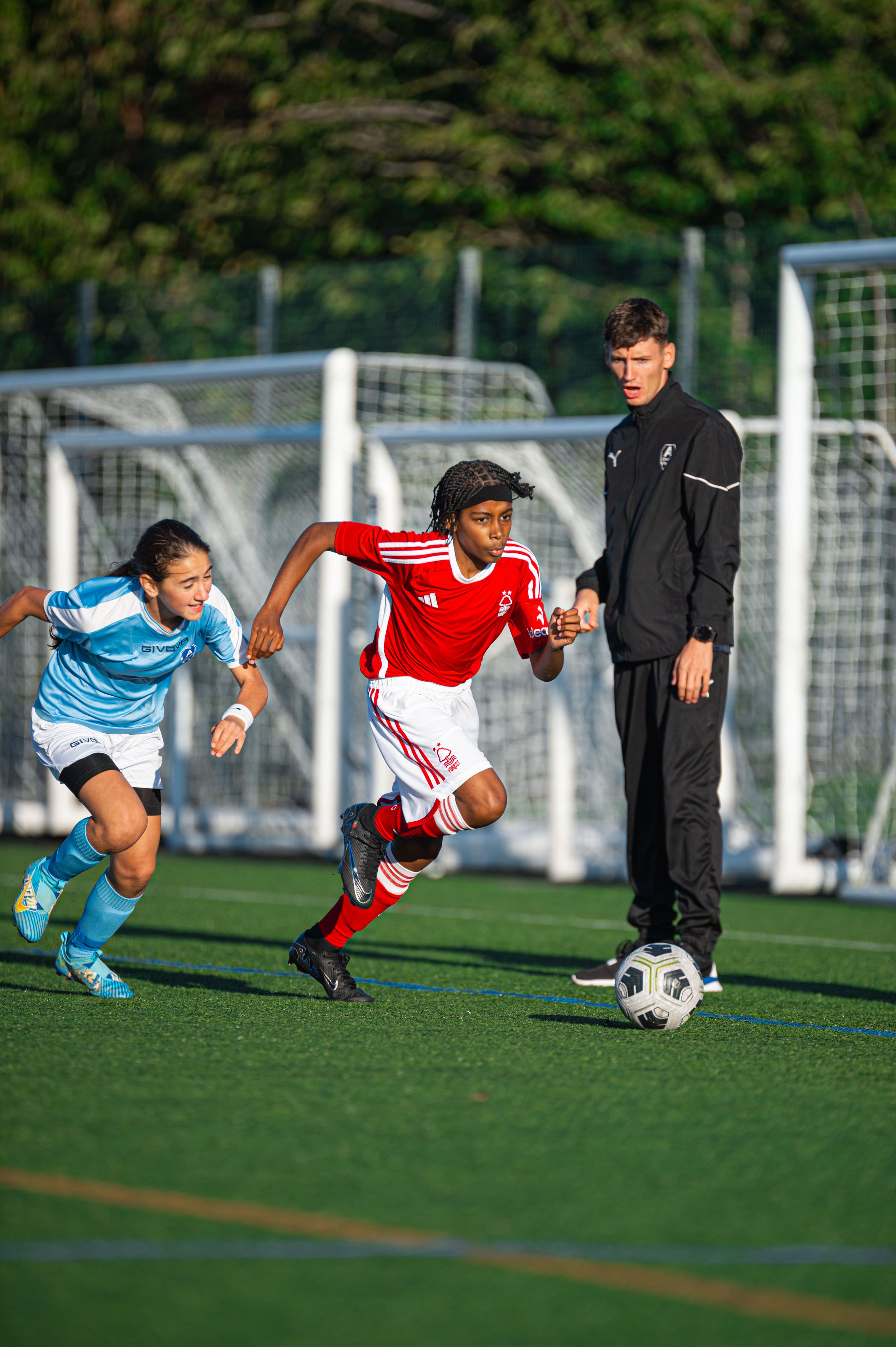 Two young female soccer players and one young male referee on a soccer field during a match. The girl in the red jersey is running towards the soccer ball, while the girl in blue is chasing behind her. The referee, dressed in black, is standing nearb