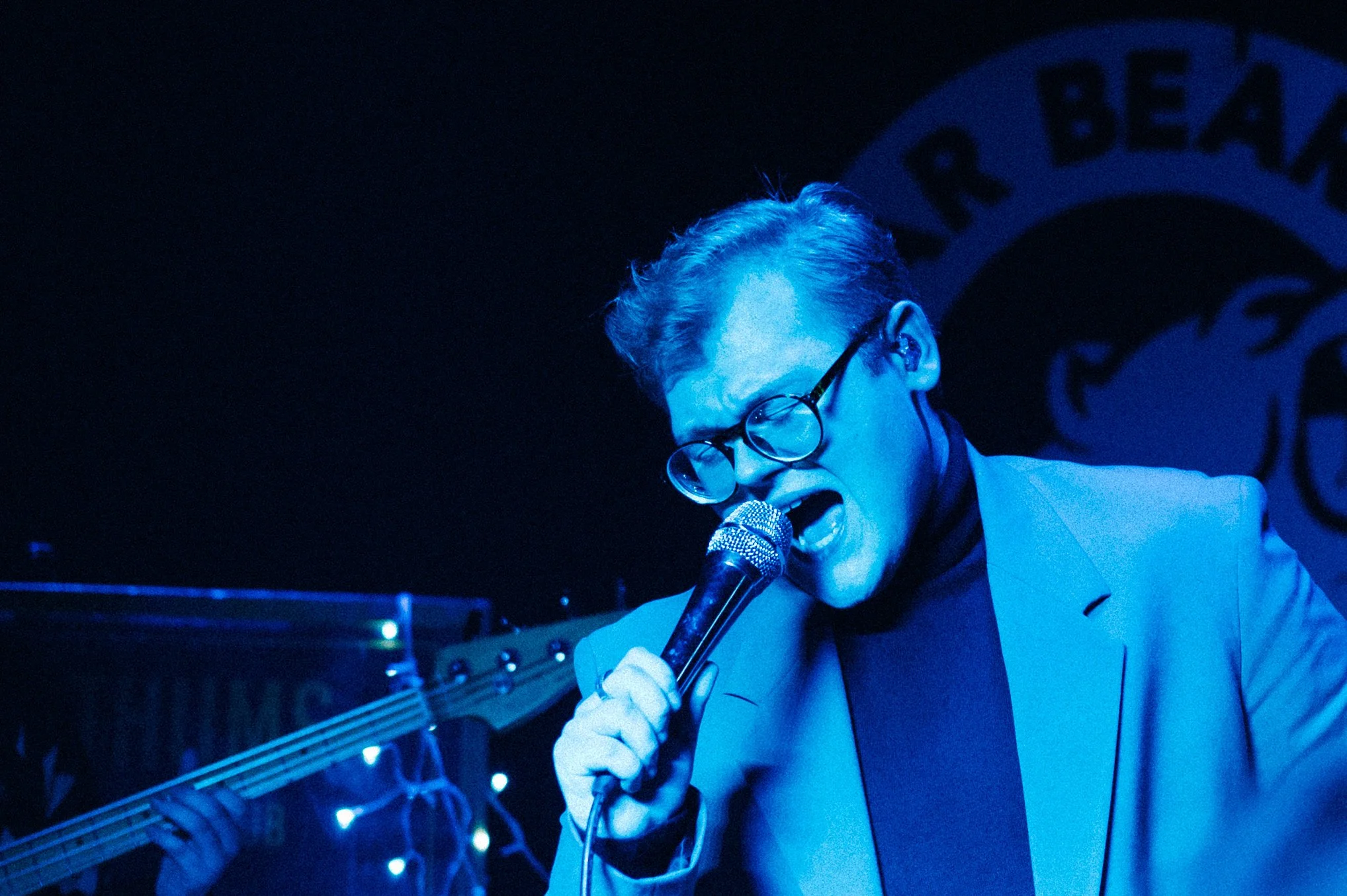 Person with glasses passionately singing or speaking into a microphone with a blue lighting atmosphere.