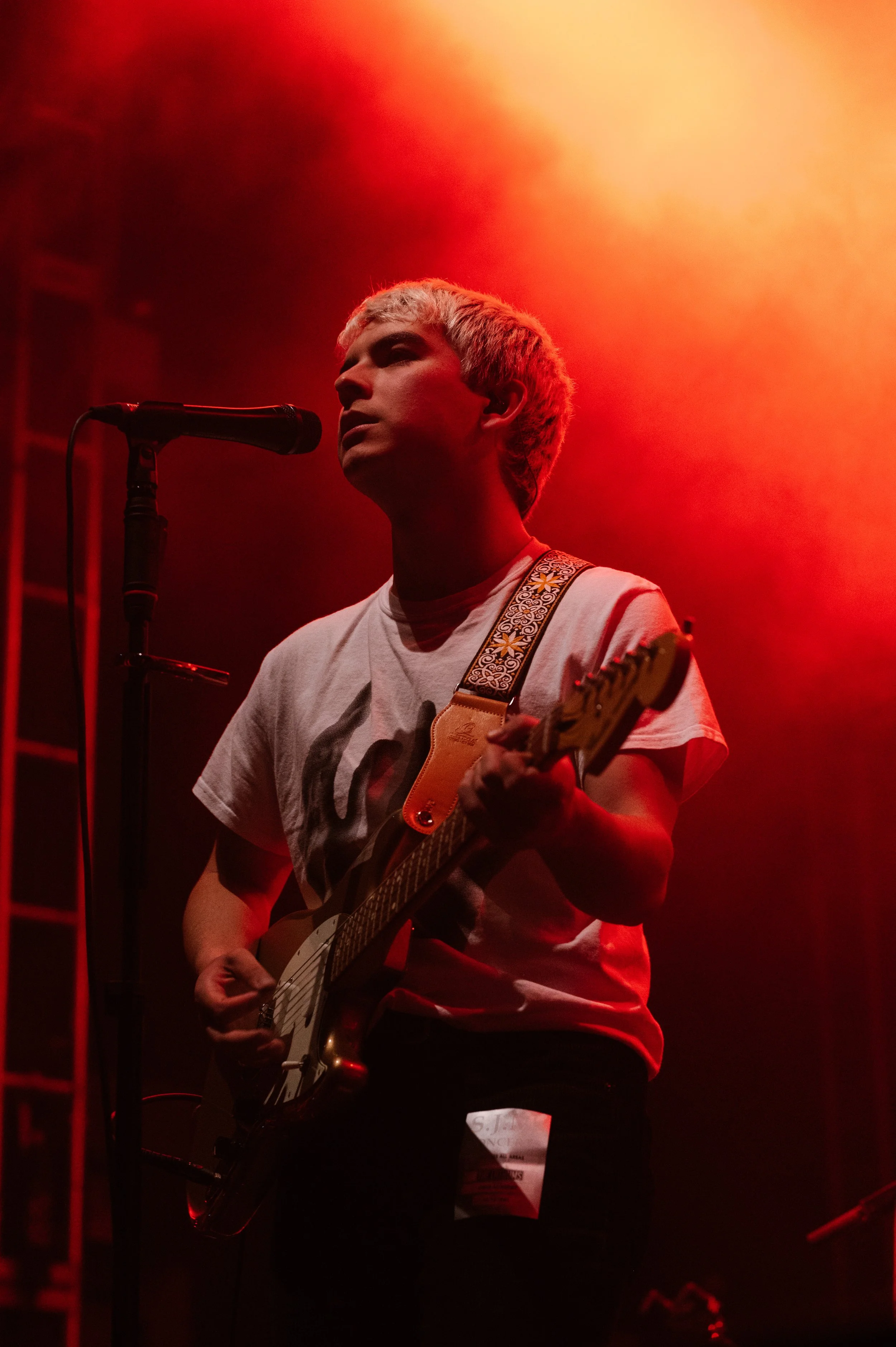 A person with short blond hair playing an electric guitar on stage, illuminated by red and orange stage lighting, with a microphone nearby.