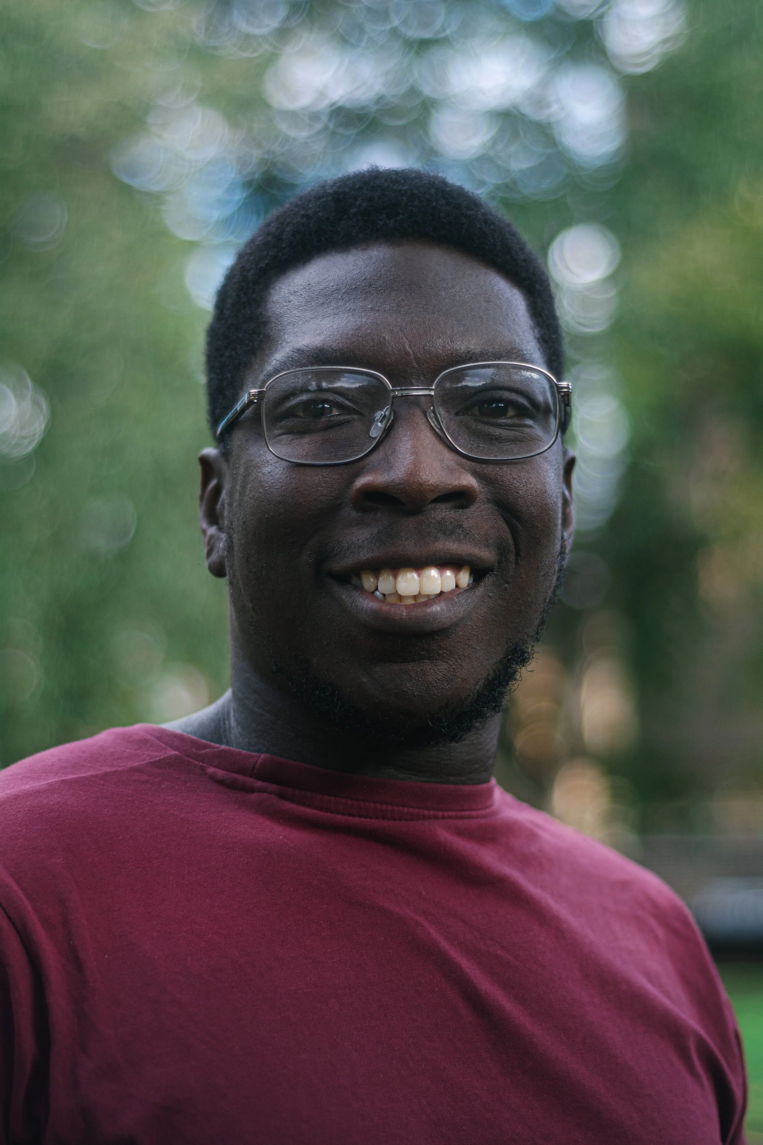 A young man with dark skin, short black hair, glasses, and a broad smile, wearing a maroon shirt, outdoors with blurred green foliage in the background.