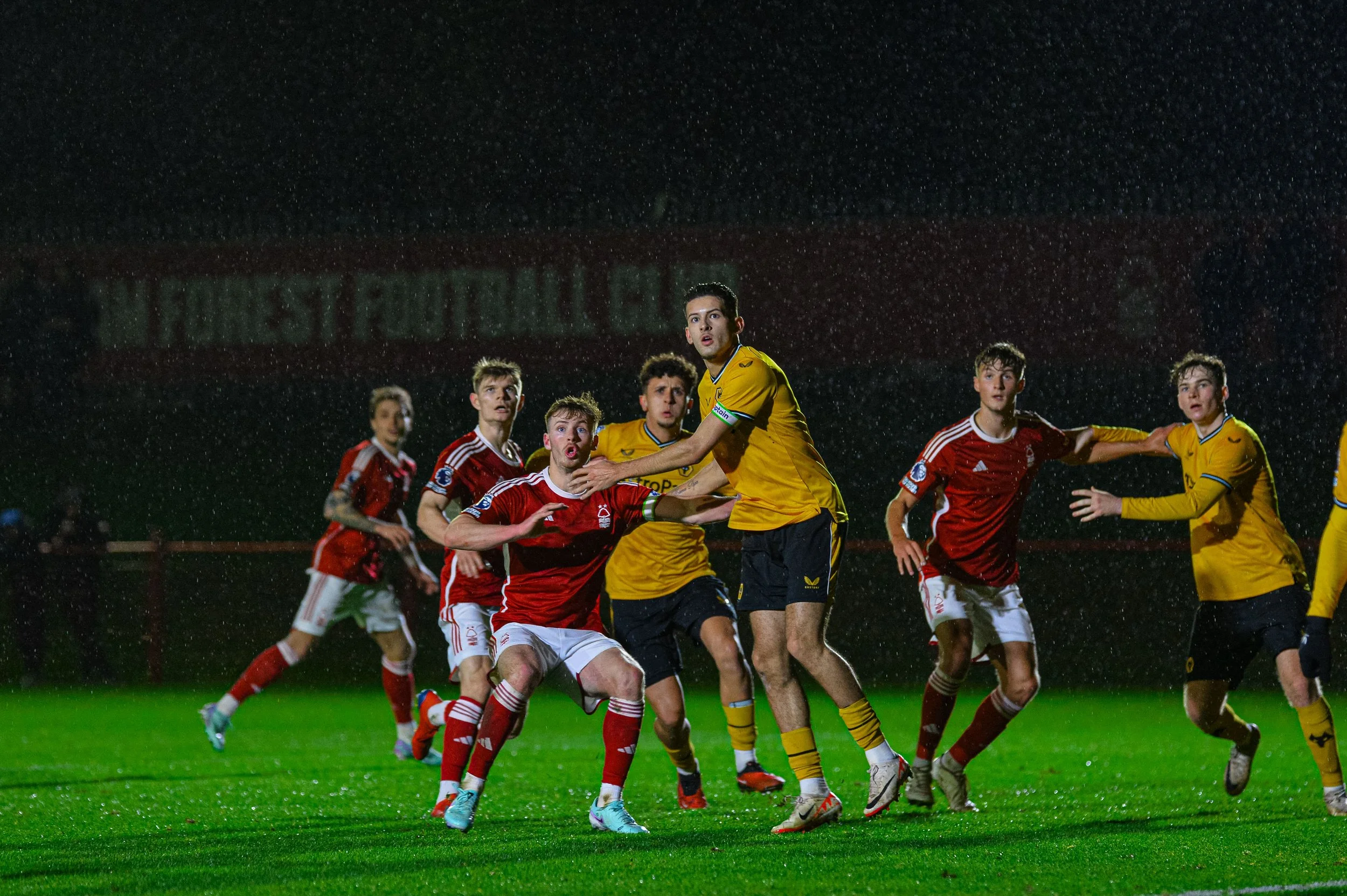 Soccer players playing in the rain during a match at night, with some players wearing red jerseys and others in yellow, on a grass field.