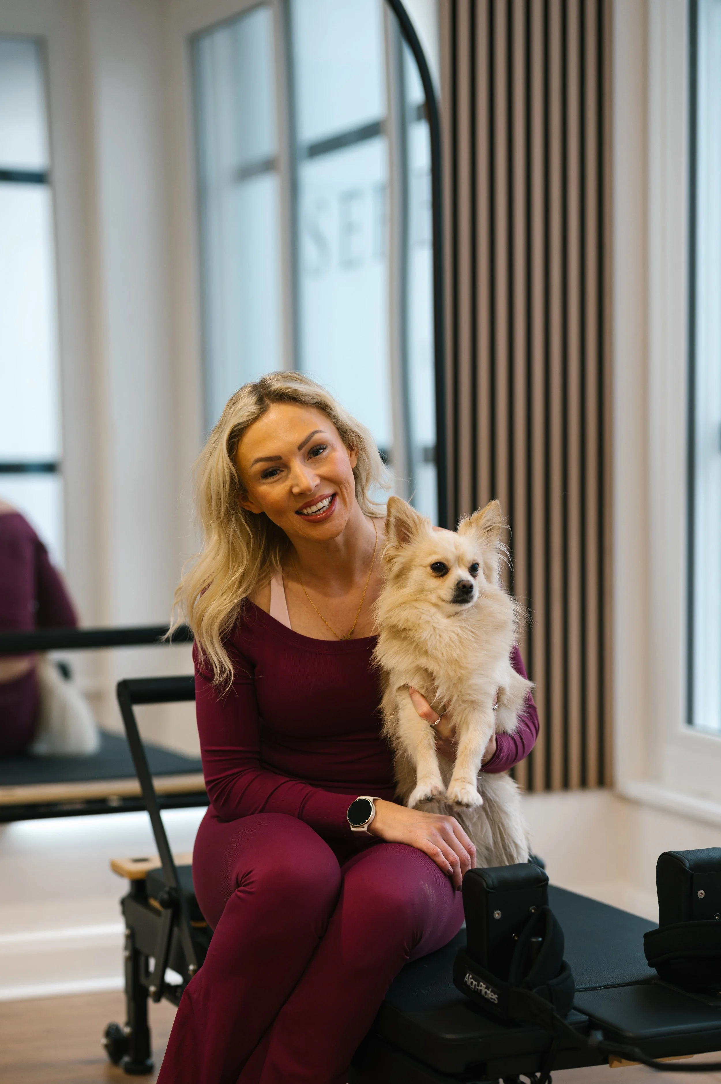 A woman with blonde hair, wearing a burgundy outfit, sitting on a Pilates reformer with a small dog in her arms. The woman is smiling and looking at the camera, with a bright studio environment and large windows in the background.