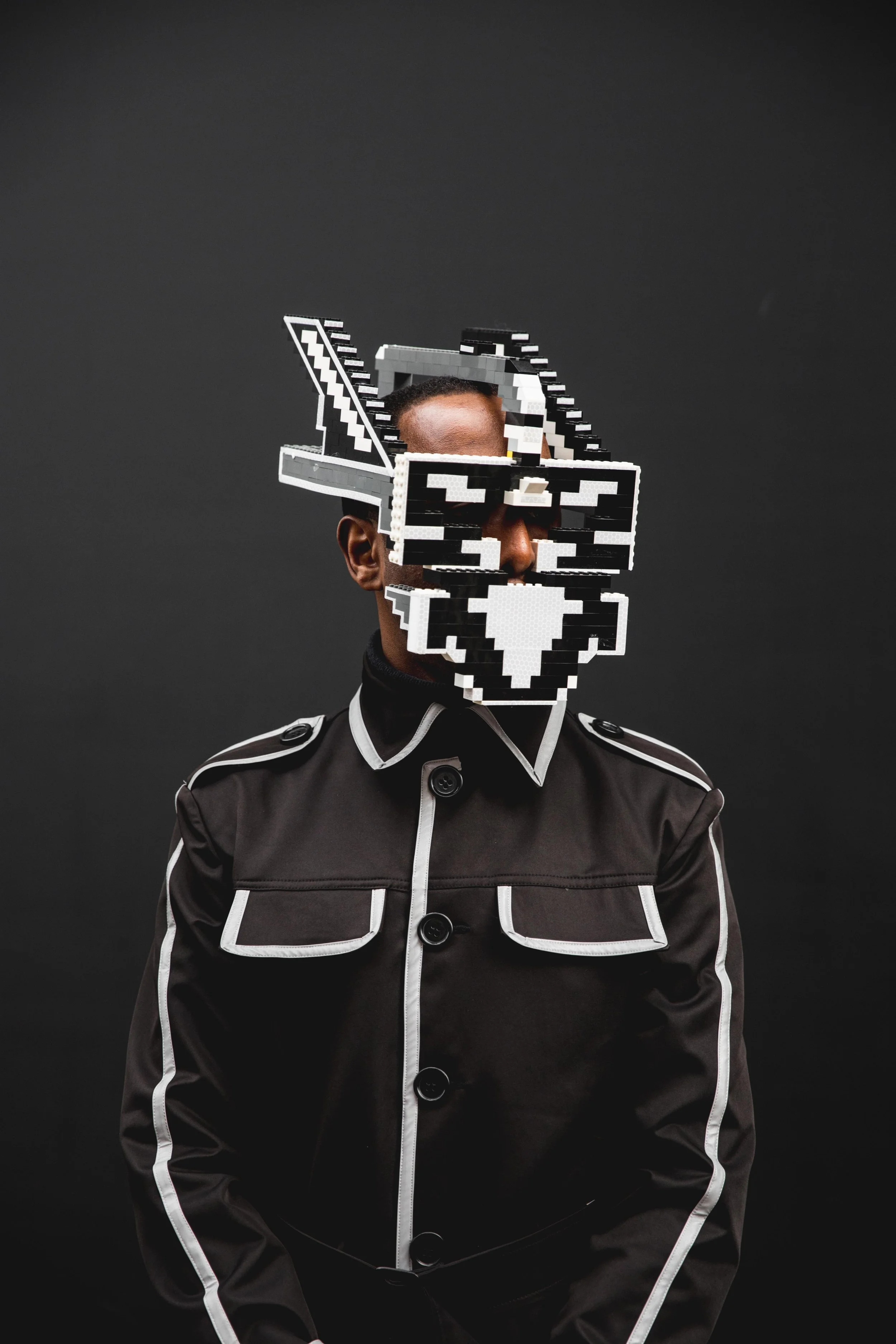 Person wearing a black jacket with white piping, standing against a dark background, with a pixelated mask covering their face.