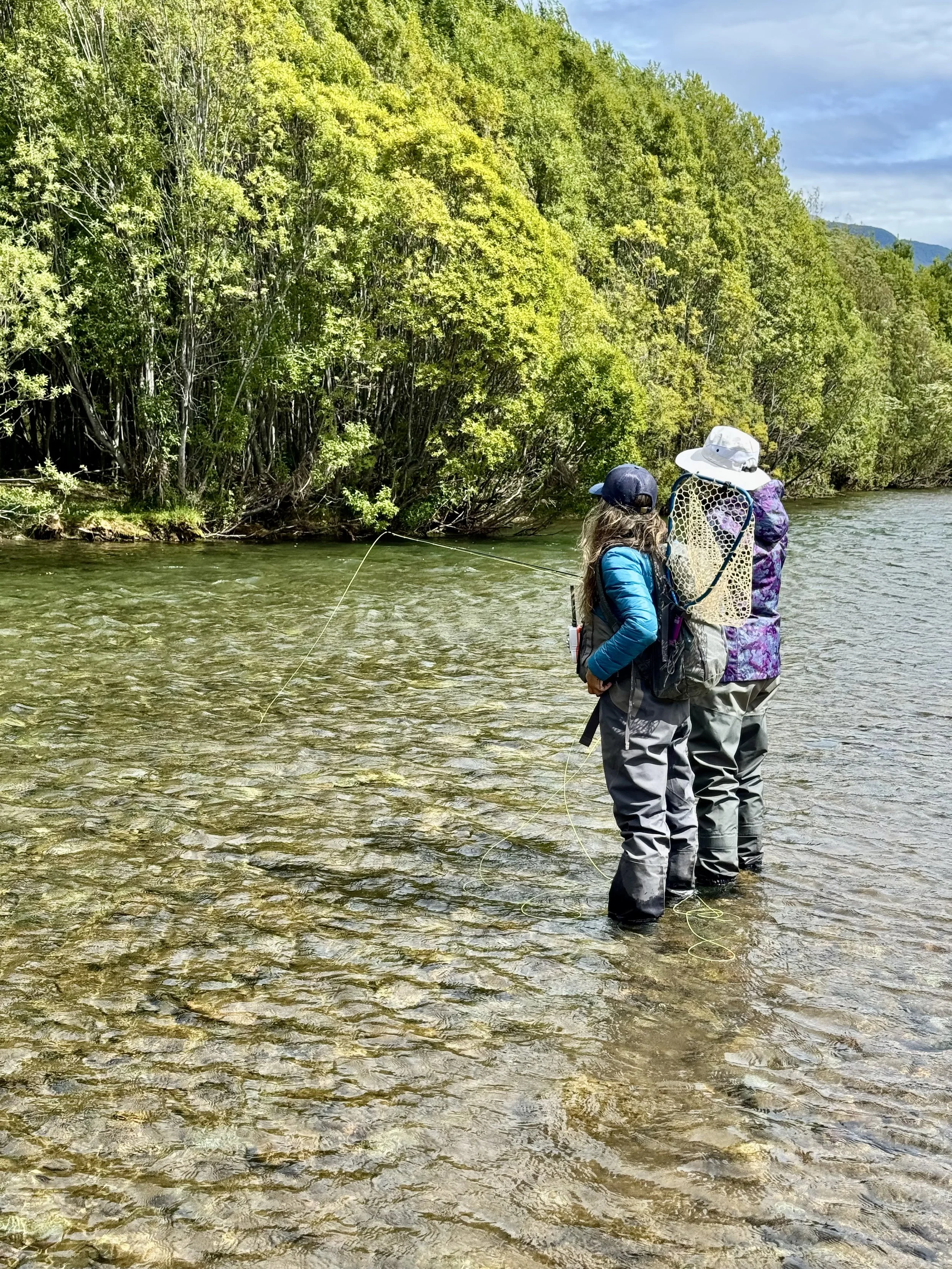 Dos personas haciendo pesca en un río rodeadas de árboles verdes bajo un cielo parcialmente nublado.