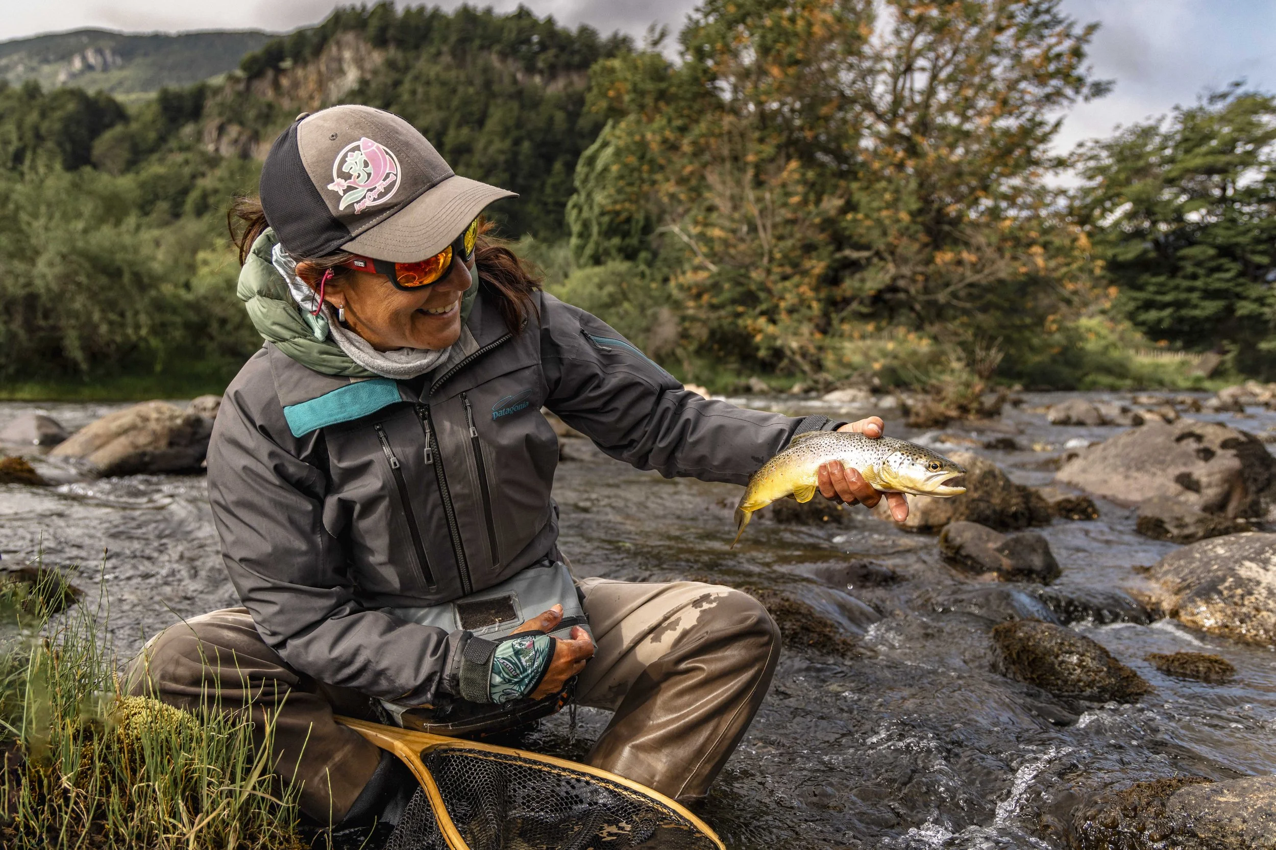 Pescadora sonriendo mientras sostiene un pez en un río rodeado de árboles y montañas.