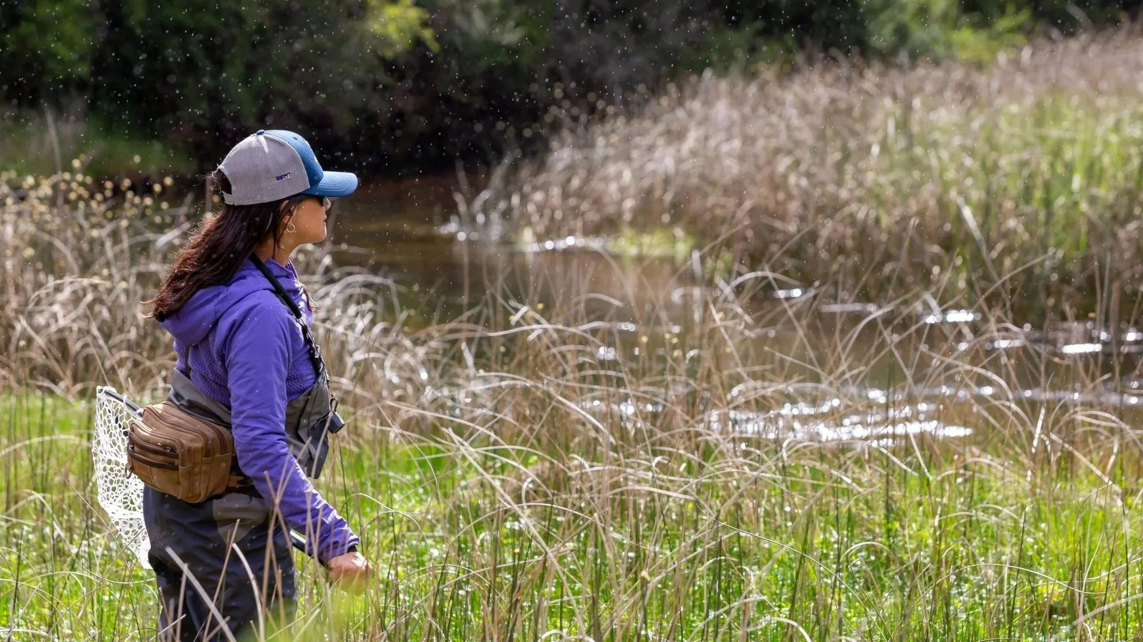 Mujer con gorra y ropa de pesca observando un cuerpo de agua rodeada de maleza en un entorno natural.
