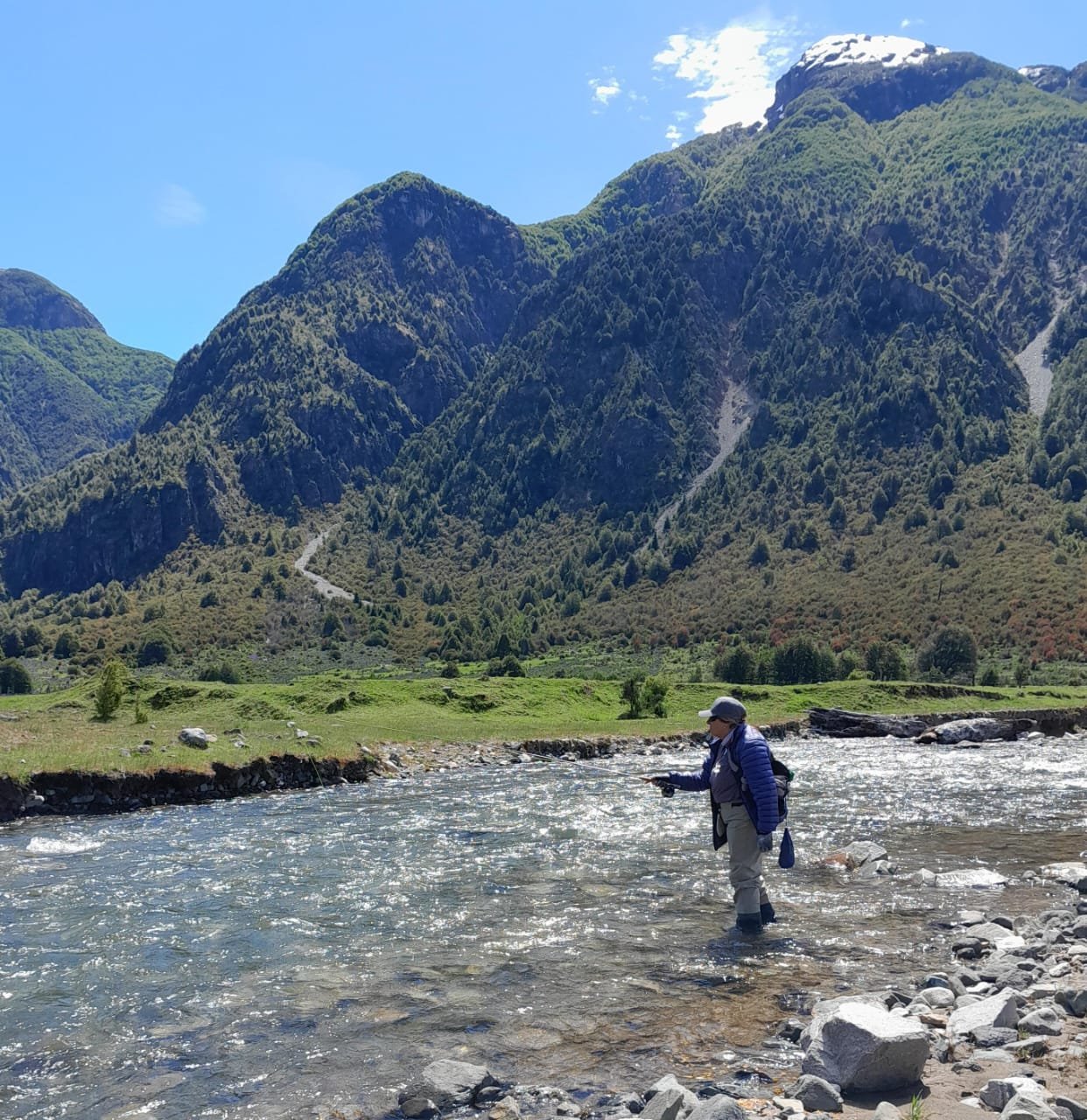 Persona pescando en río con montañas verdes y cielo azul al fondo.