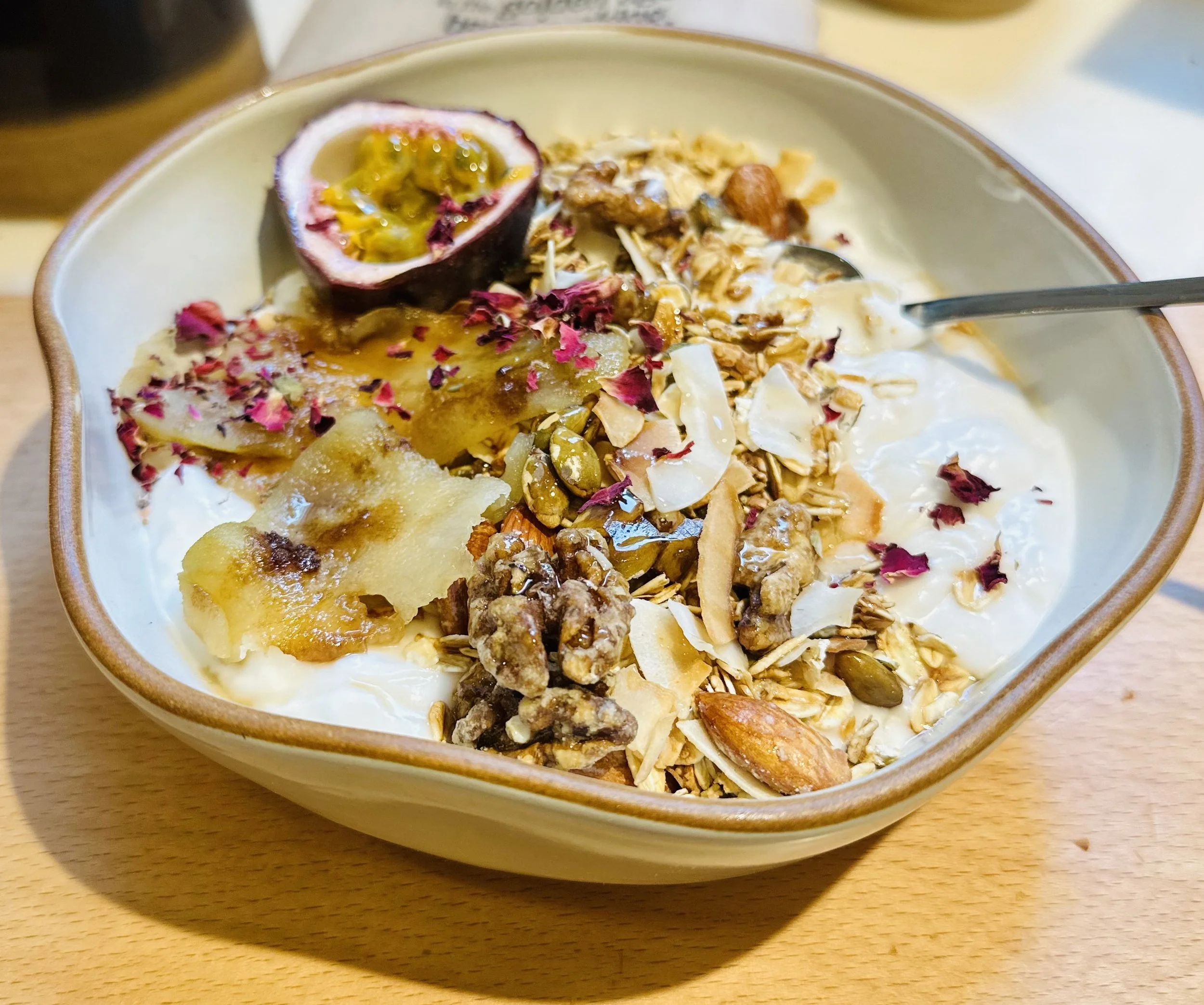 A bowl of yogurt topped with granola, nuts, dried rose petals, and a halved passion fruit with yellow pulp.