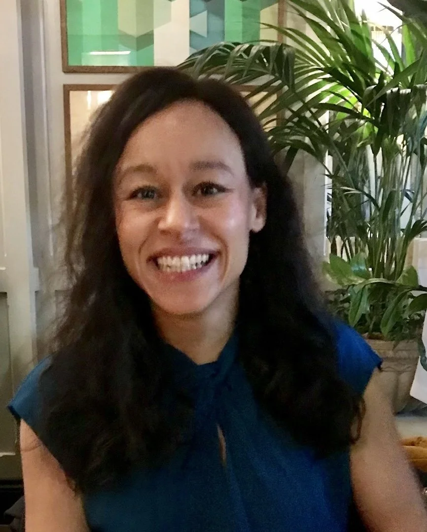 Smiling woman with long dark hair and a blue top, sitting indoors near green plants.