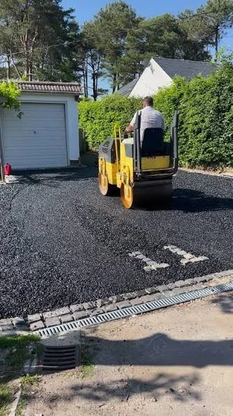 A person operating a small yellow road roller on freshly paved asphalt in a residential driveway.