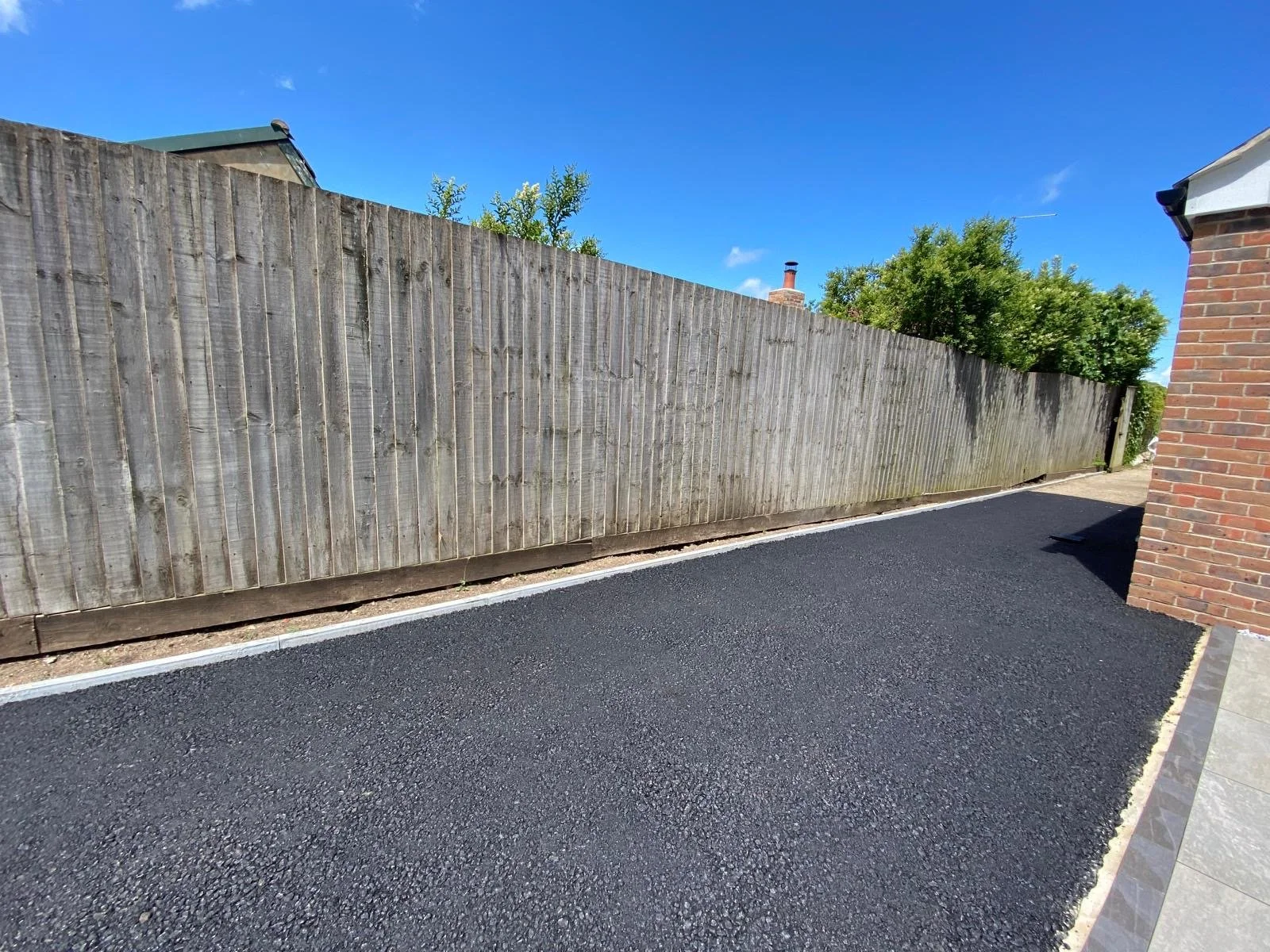 A paved driveway next to a brick house and a tall wooden fence, with trees and a blue sky in the background.