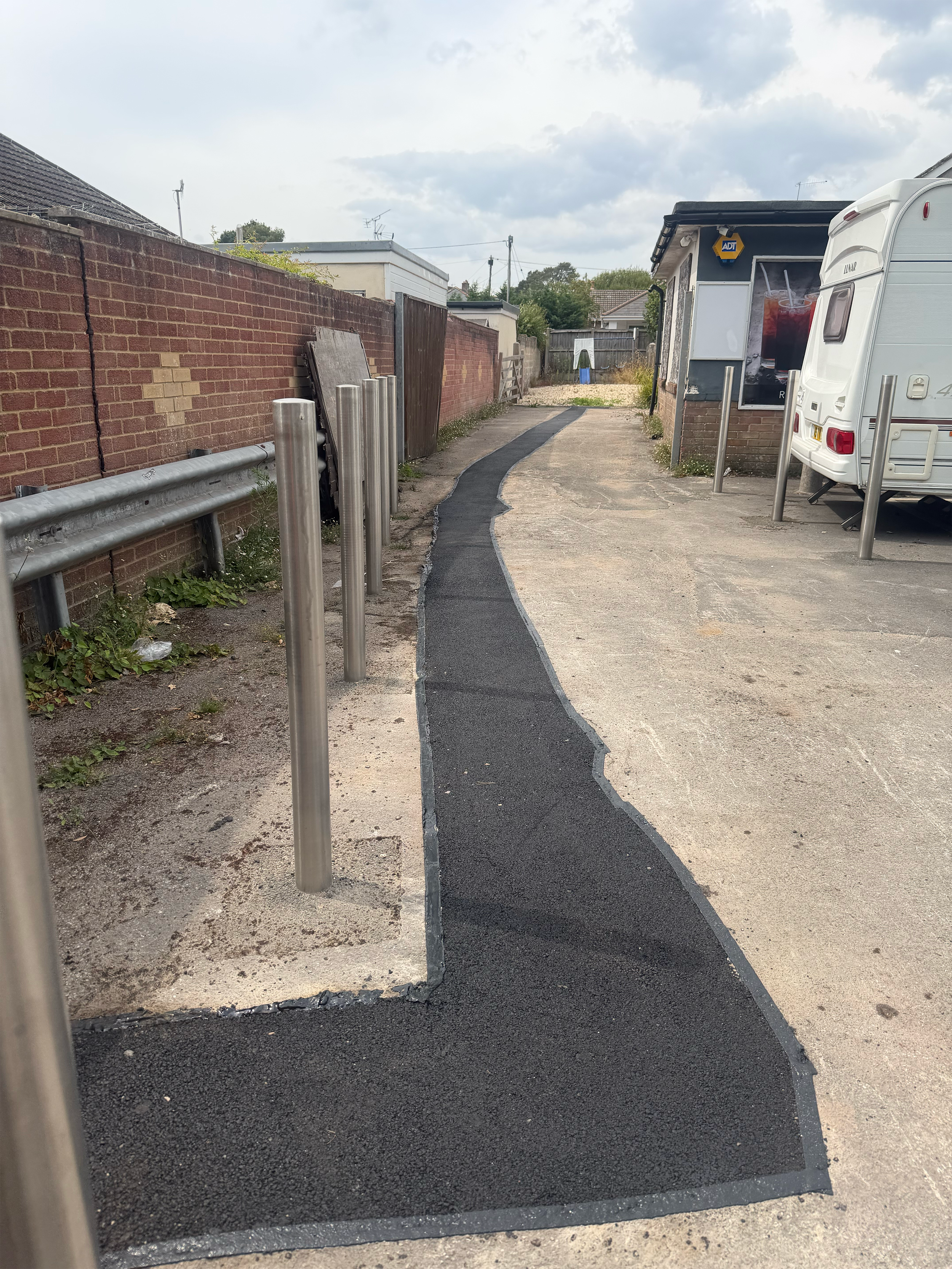 A narrow black asphalt path running through an outdoor area, bordered by a brick wall on the left and a concrete parking lot on the right. The path curves slightly to the right.