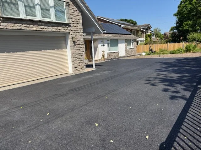 Freshly paved asphalt driveway next to a house with stone and white siding, a garage door, and solar panels on the roof. There are some leaves on the driveway and a wooden fence in the background.
