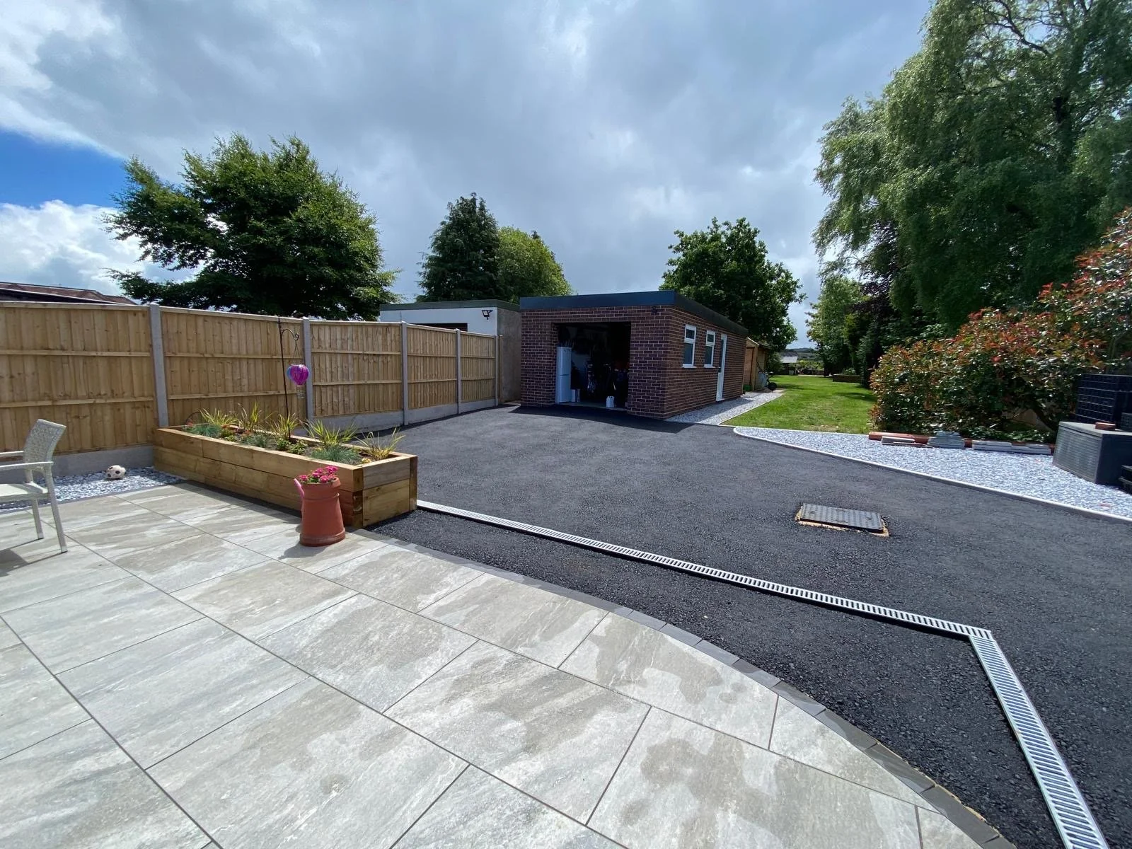 A backyard with a paved patio area, a flower bed, a wooden fence, a small brick shed, and a grassy lawn with trees and shrubs under a partly cloudy sky.