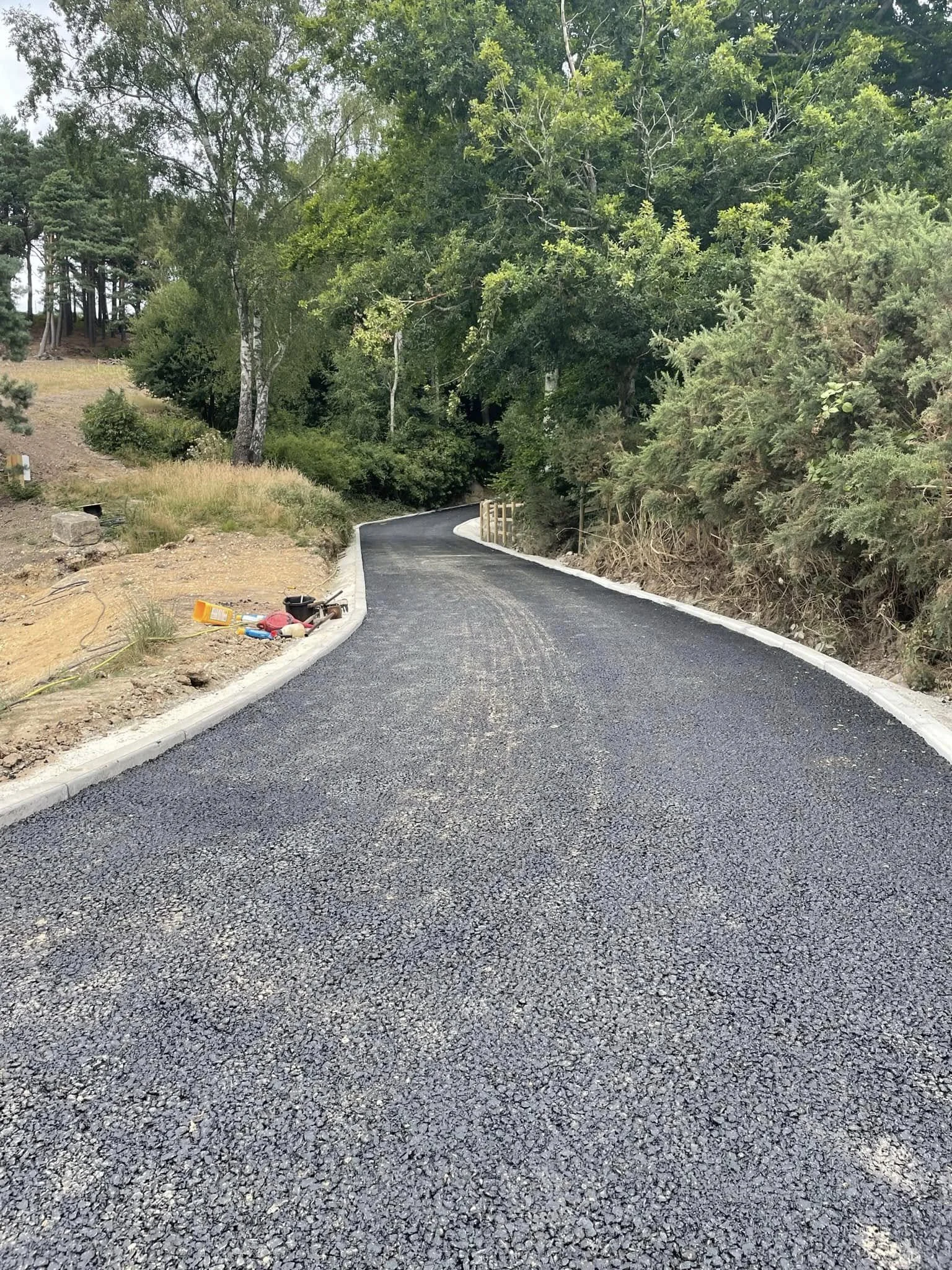 A newly paved black asphalt road winding through a wooded area with green trees on both sides, some construction tools and materials placed on the roadside.