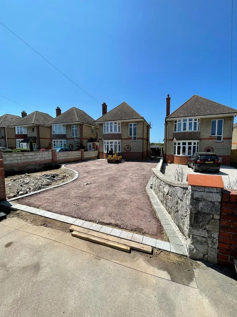 Residential driveway construction with gravel and paving stones, uneven ground, and a small construction vehicle parked near the house, under a clear blue sky.