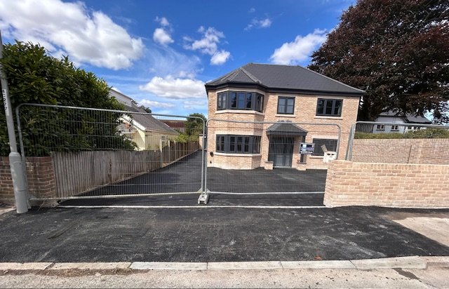 Newly paved driveway in front of a two-story brick house with a black roof, black windows, and a small porch, surrounded by a brick wall and metal fencing.
