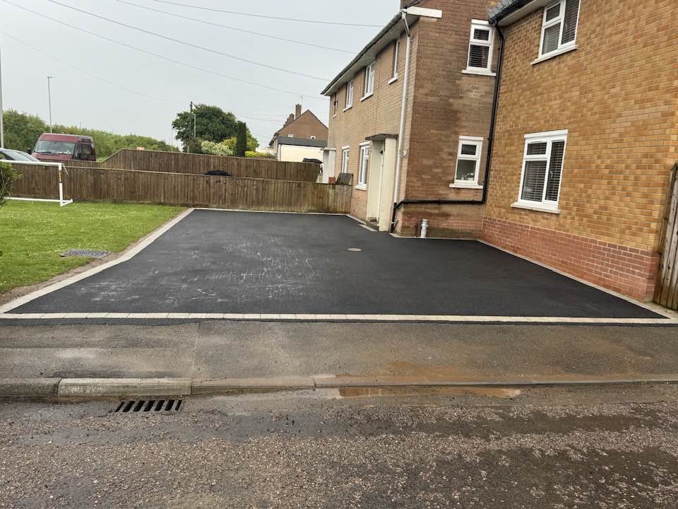 Finished freshly paved black asphalt driveway in front of a brick house, bordered by a concrete edge, with a small grass lawn to the left and a wooden fence at the back.
