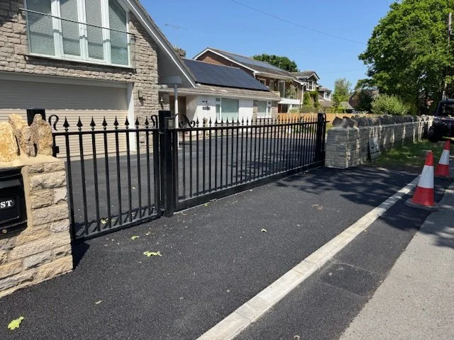 A black metal gate and stone pillars blocking a driveway in front of a residential house with solar panels on the roof and trees in the background.