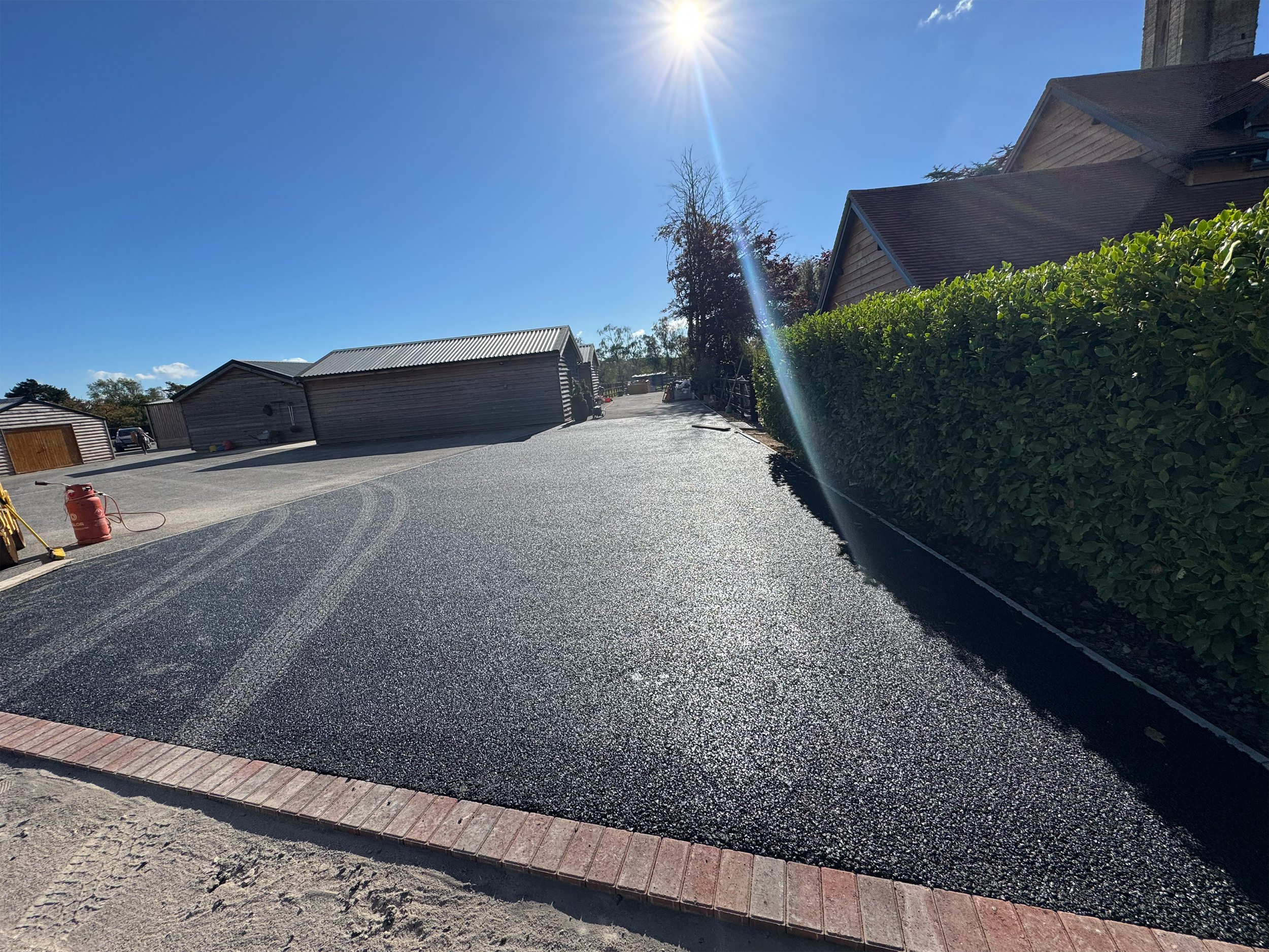 Construction site with freshly paved asphalt, brick border, and green hedges along the sidewalk. Buildings and blue sky with the sun in the sky are visible.