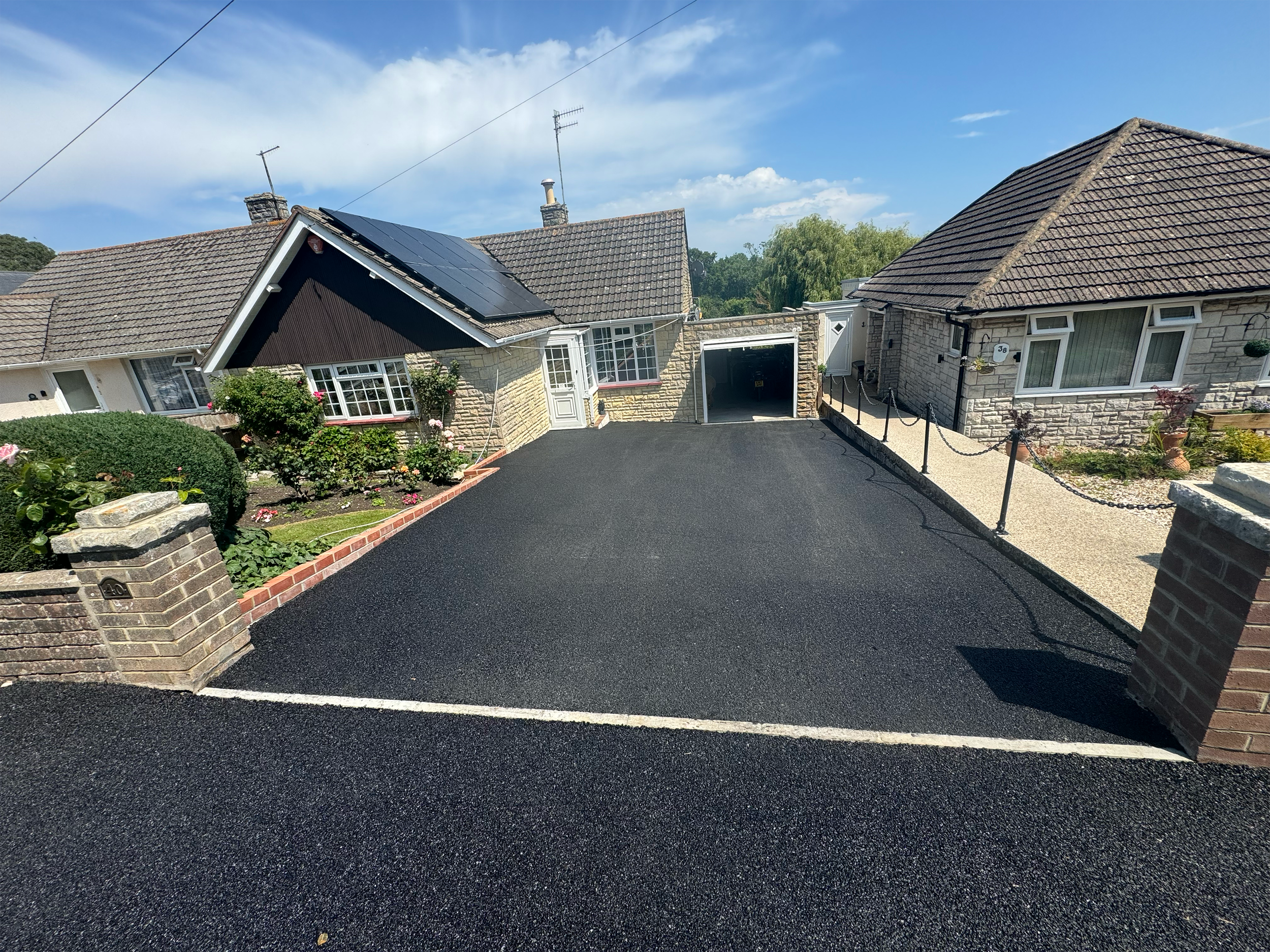 A residential street with two houses, one with solar panels on the roof, and a driveway with freshly paved black asphalt. The sky is clear with some clouds.