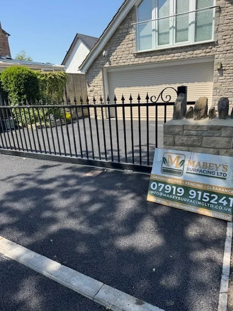 A black metal fence in front of a house with a stone exterior and large window. There is a sign advertising Mabey's Surfacing Ltd with a phone number and website, placed on the asphalt driveway.
