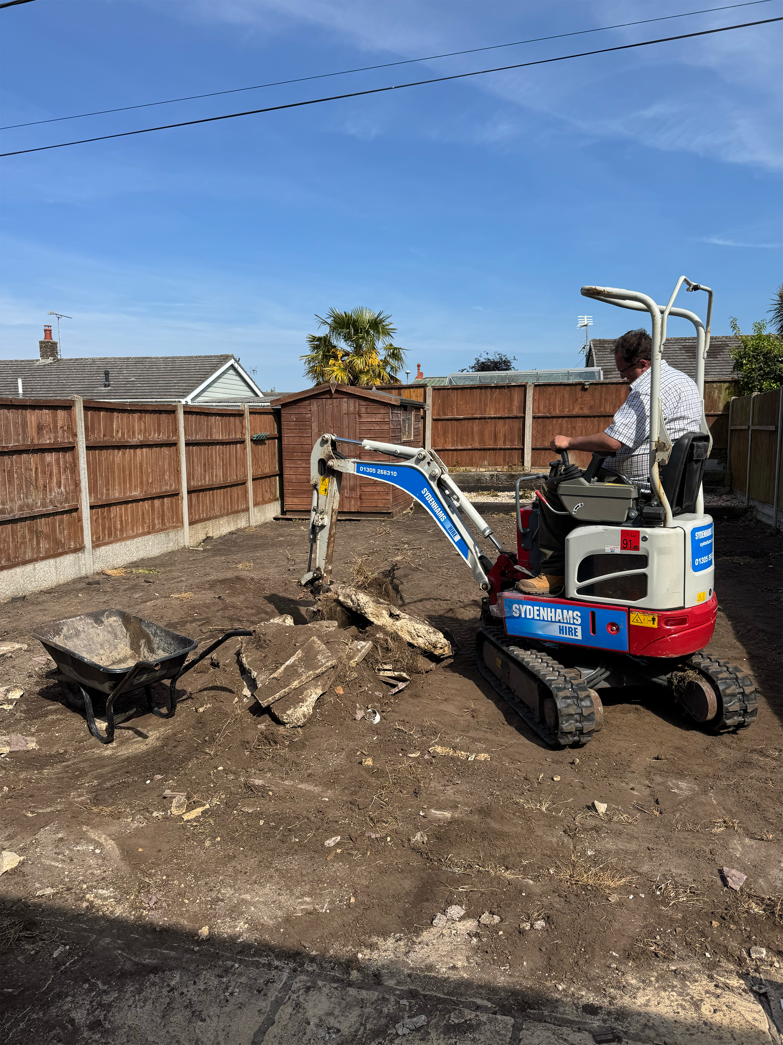 A man operating a small excavator in a backyard, with a wooden fence, a small garden shed, and a few trees in the background. The ground appears to be under construction or renovation.