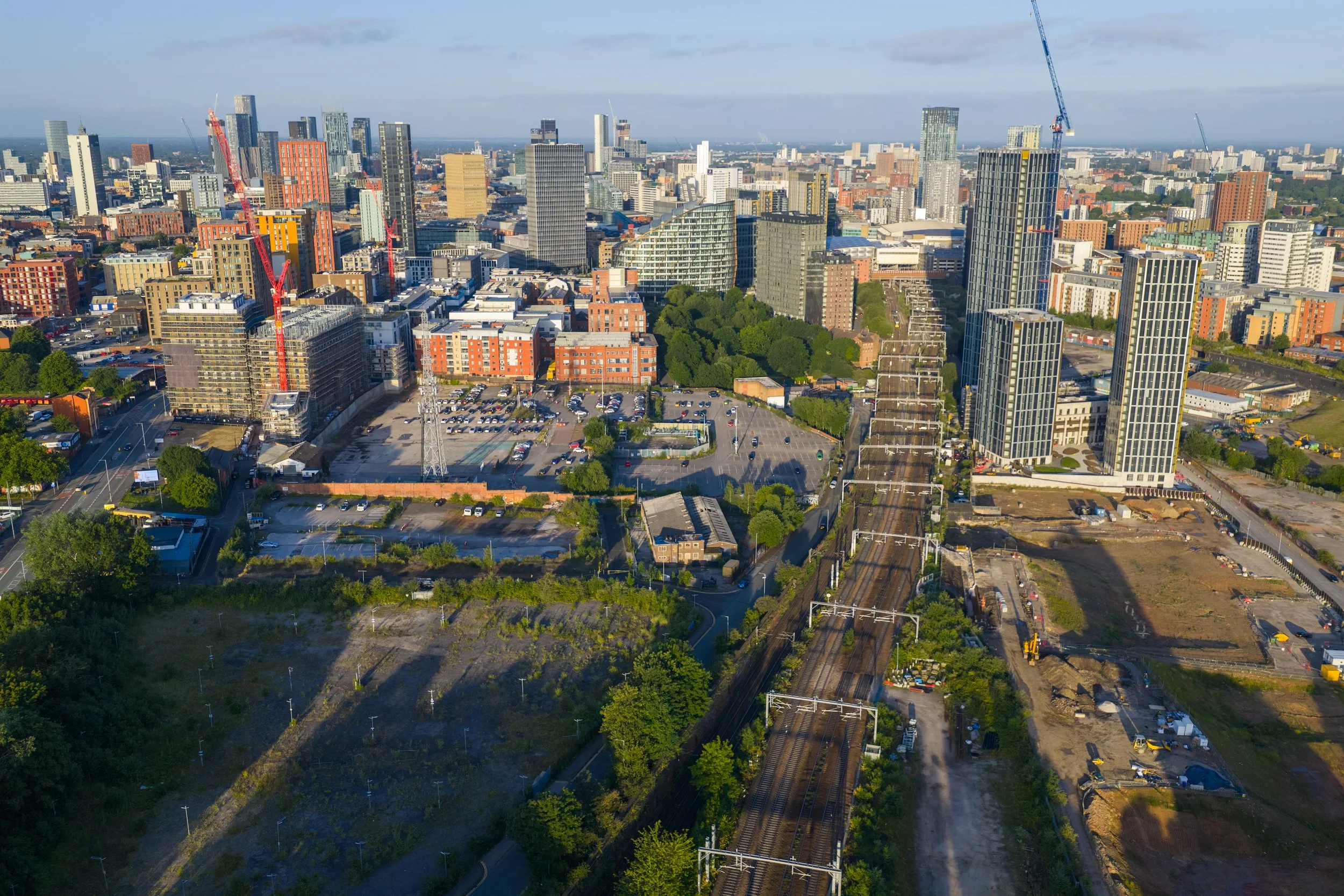 A city skyline with tall skyscrapers under a clear sky, construction cranes, and a railway track in the foreground.