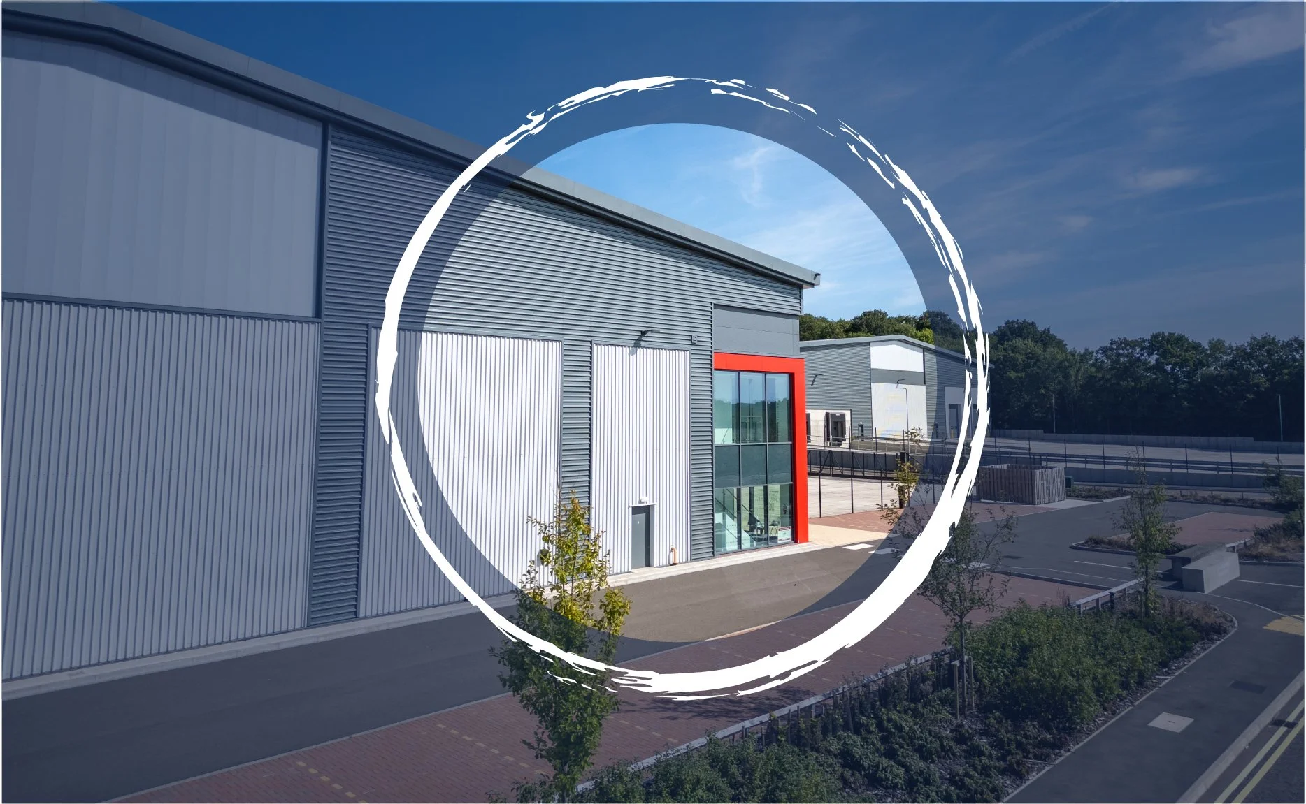 Exterior of modern industrial building with metal siding, large windows, and a red accent frame, surrounded by a paved parking area and small trees.