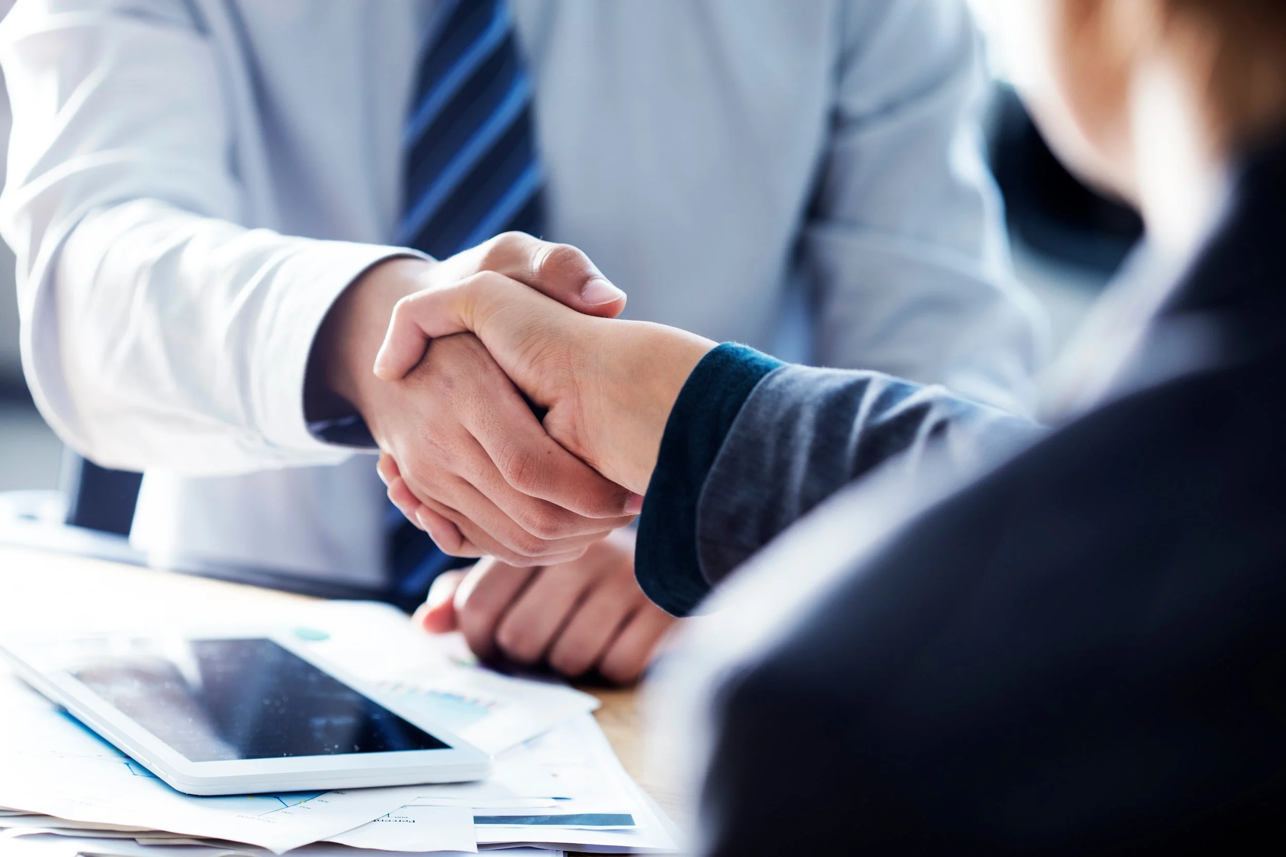 Two individuals shaking hands in a professional setting, with documents and a tablet on the table.
