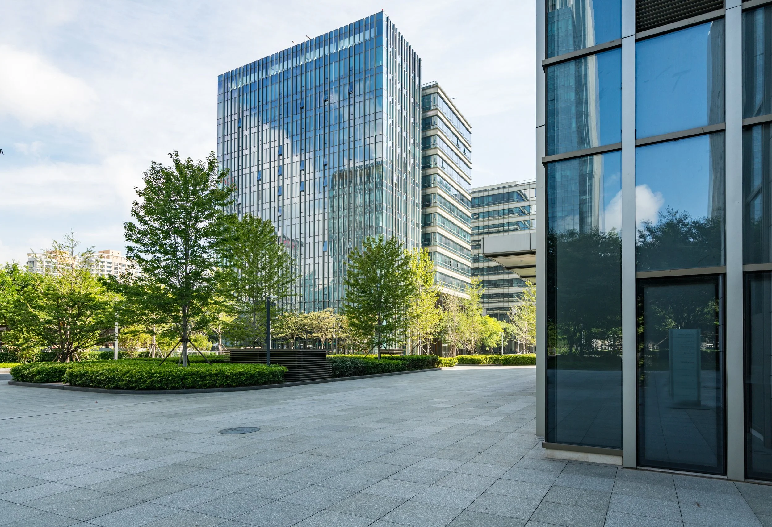 Modern office buildings with glass facades, surrounded by green trees and a paved plaza area.