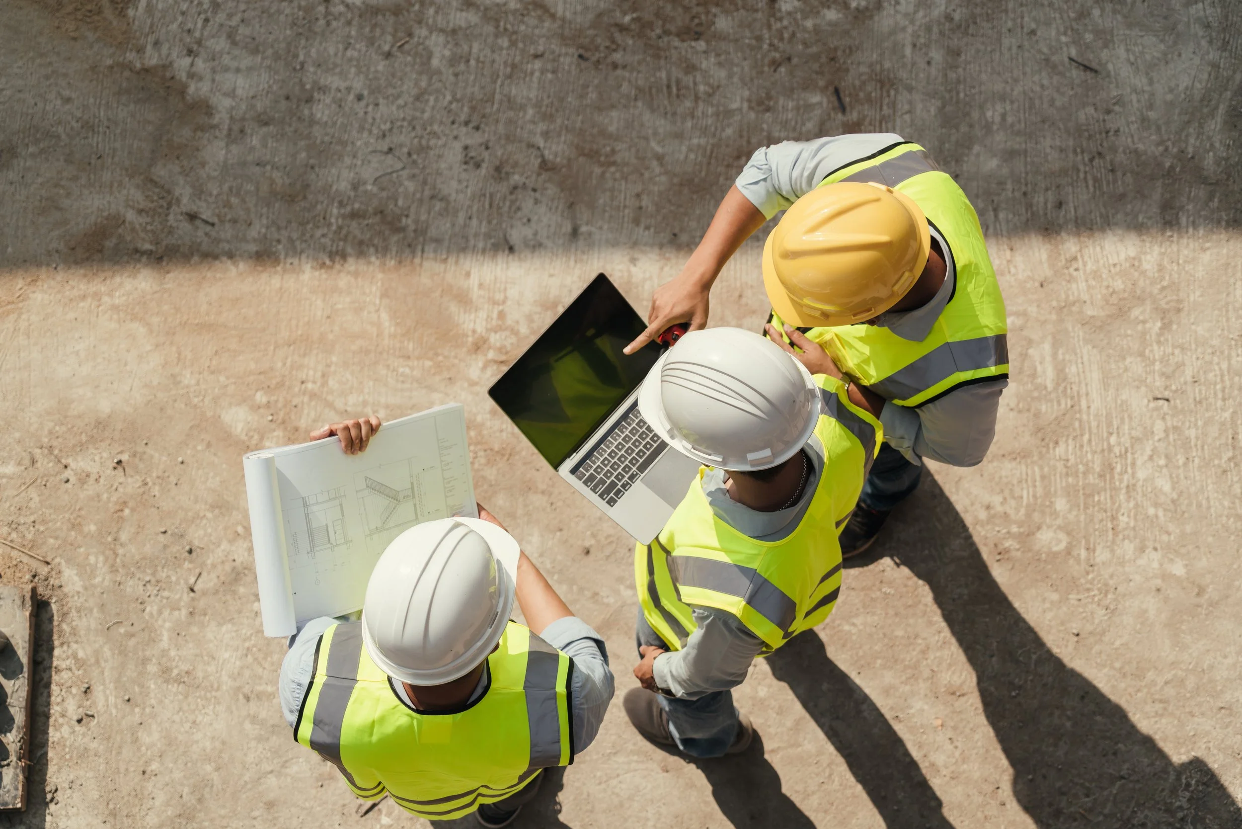 Construction workers wearing yellow safety vests and hard hats discussing blueprints on a construction site.