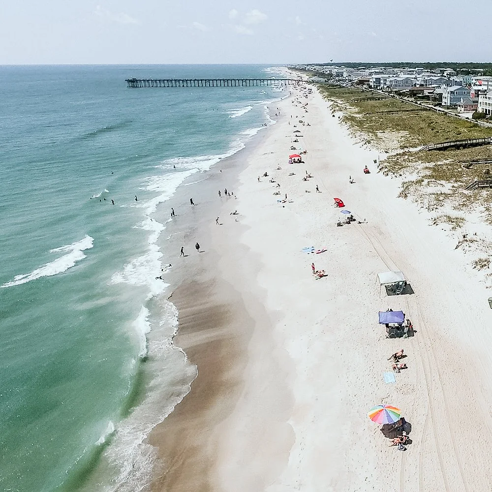 Bird's-eye view of a sandy beach with people swimming, sunbathing, and under umbrellas, adjacent to a row of houses and a pier extending into the ocean.