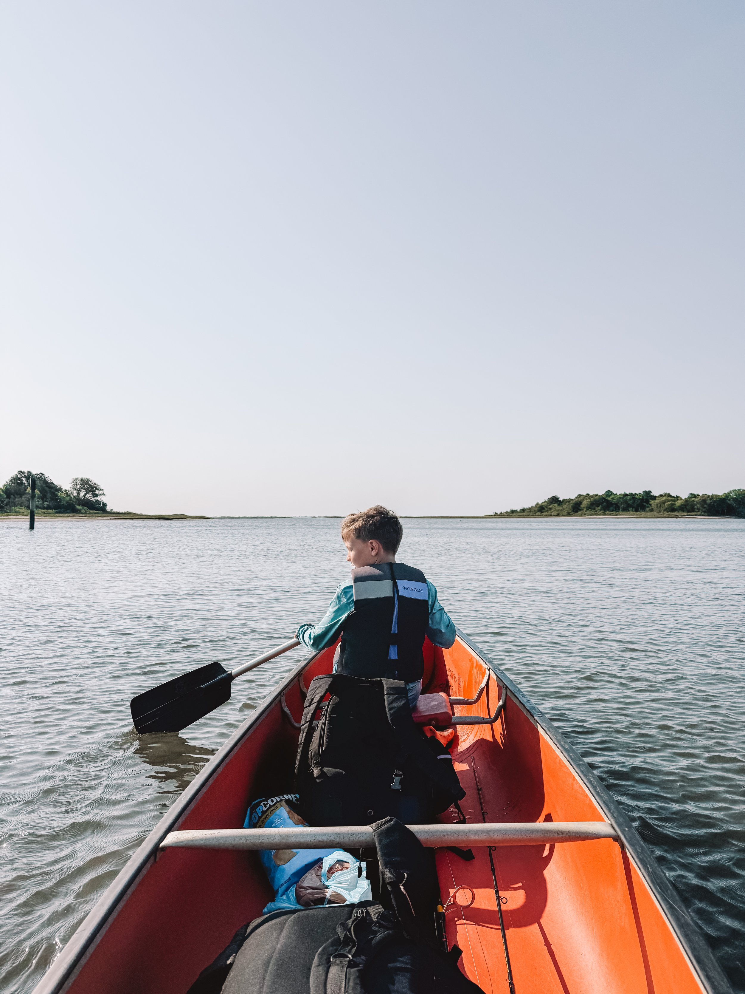 A boy sitting in a red canoe on a calm body of water with a landscape of trees and hills in the background, paddling with a black oar on a clear day.