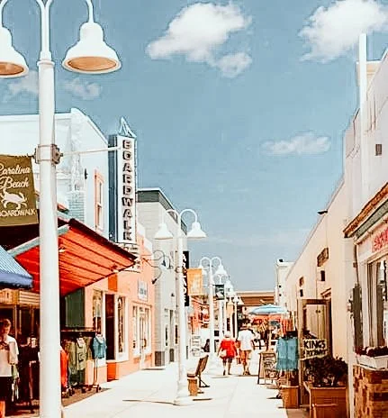A lively street scene at a shopping or entertainment district with storefronts, streetlights, and people walking, under a blue sky with clouds.