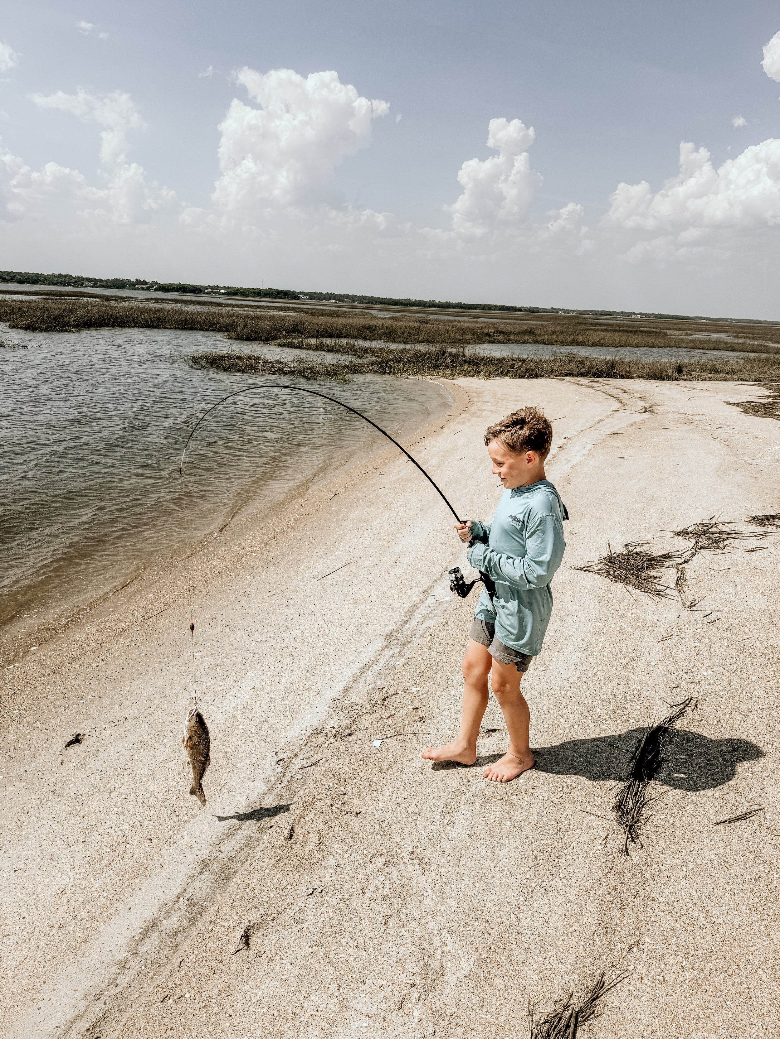 A young boy on a sandy beach, barefoot, holding a fishing rod with a fish attached, looking at it with a smile, with grasses and water in the background under a cloudy sky.
