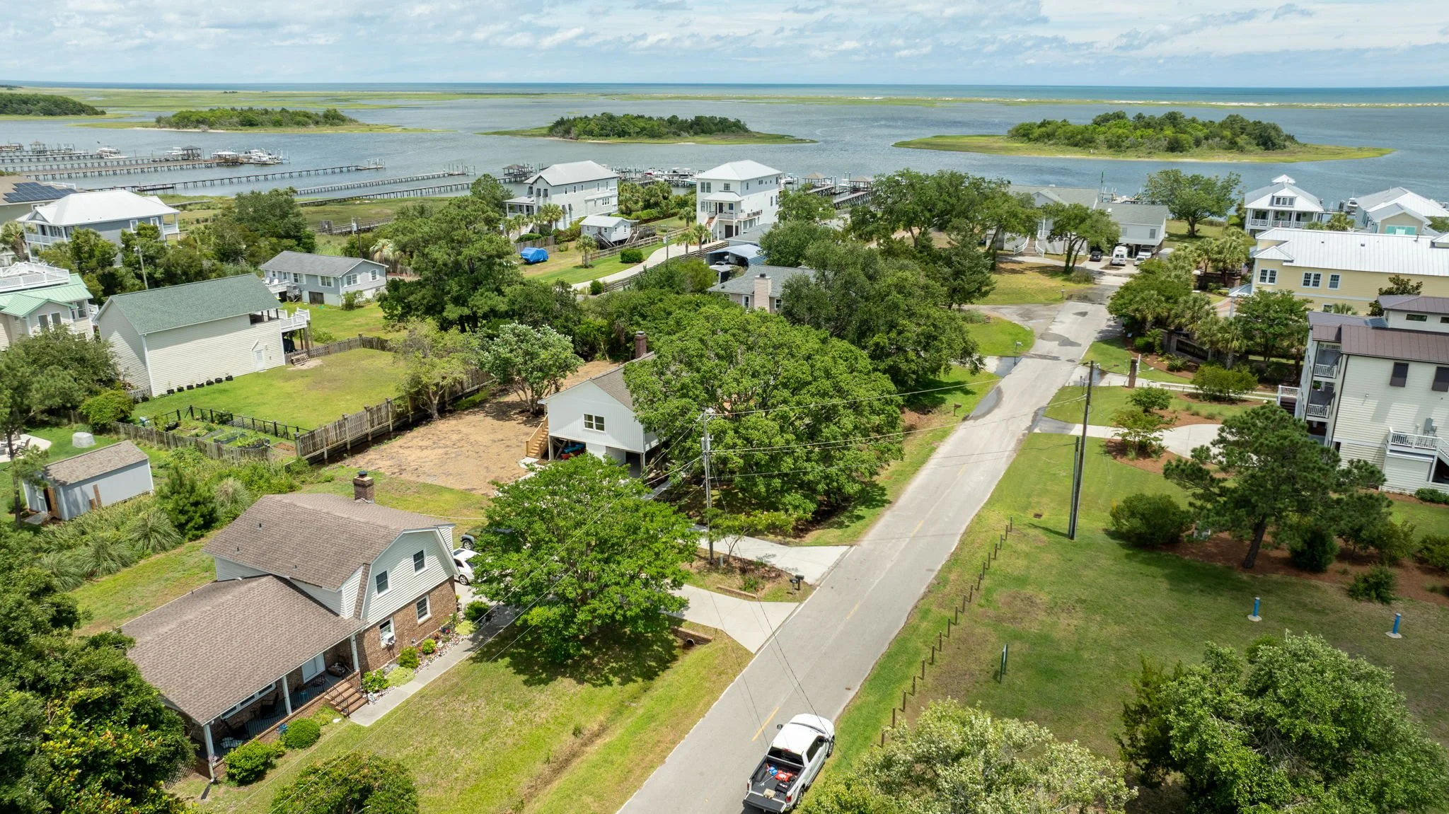 Aerial view of a coastal neighborhood with houses, trees, a road, and a large body of water with small islands and boat docks in the background.