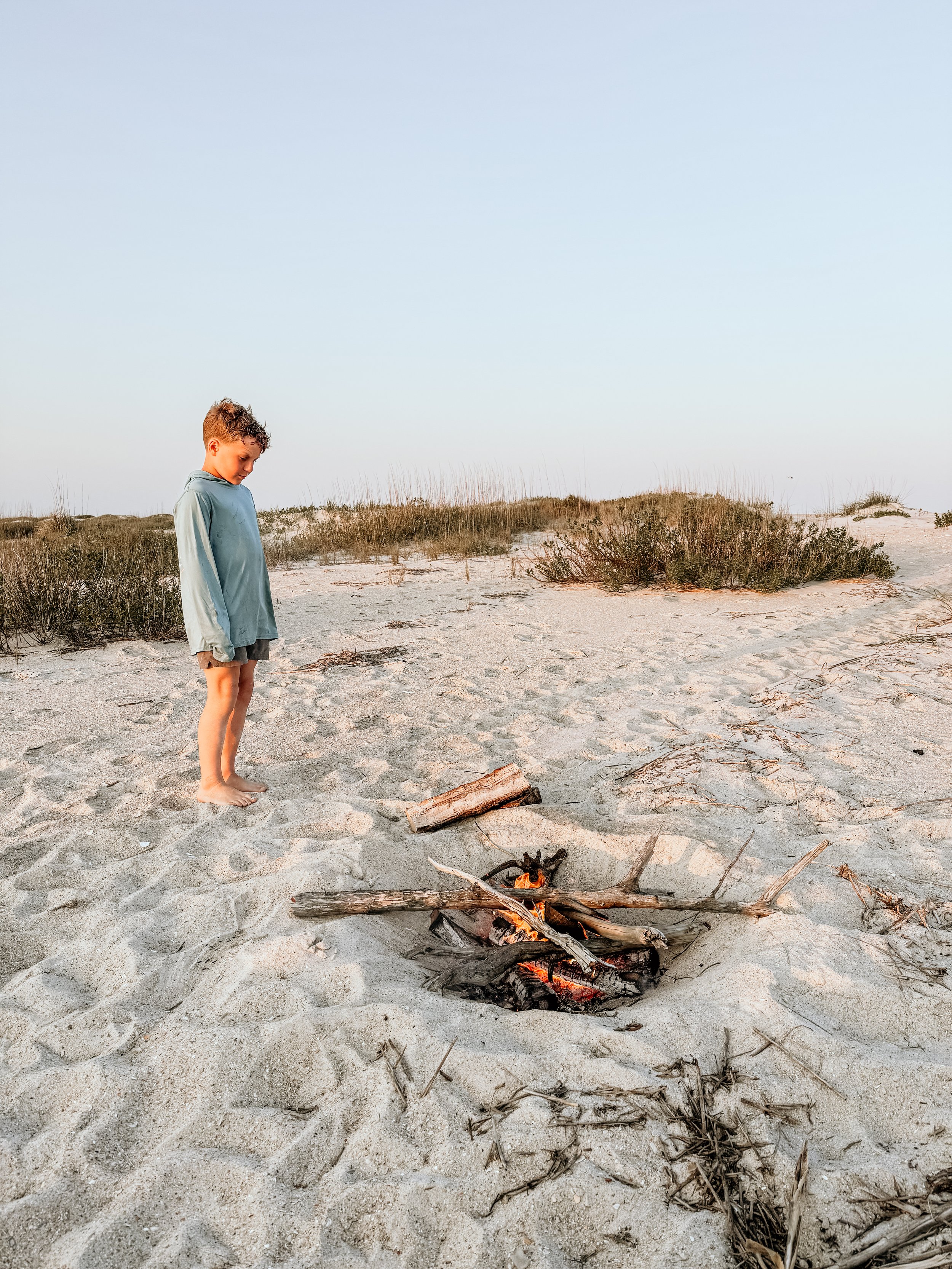 Child standing on sandy beach near small campfire made of logs with dry grass and shrubs in the background under clear sky.