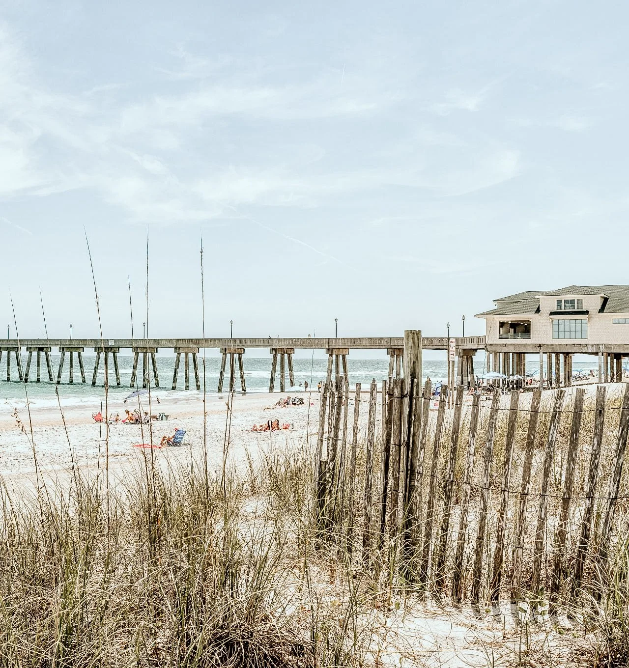 Beach scene with a pier extending over the ocean, a beach house on stilts, and beachgoers relaxing on the sand, viewed through a fence and tall grass.