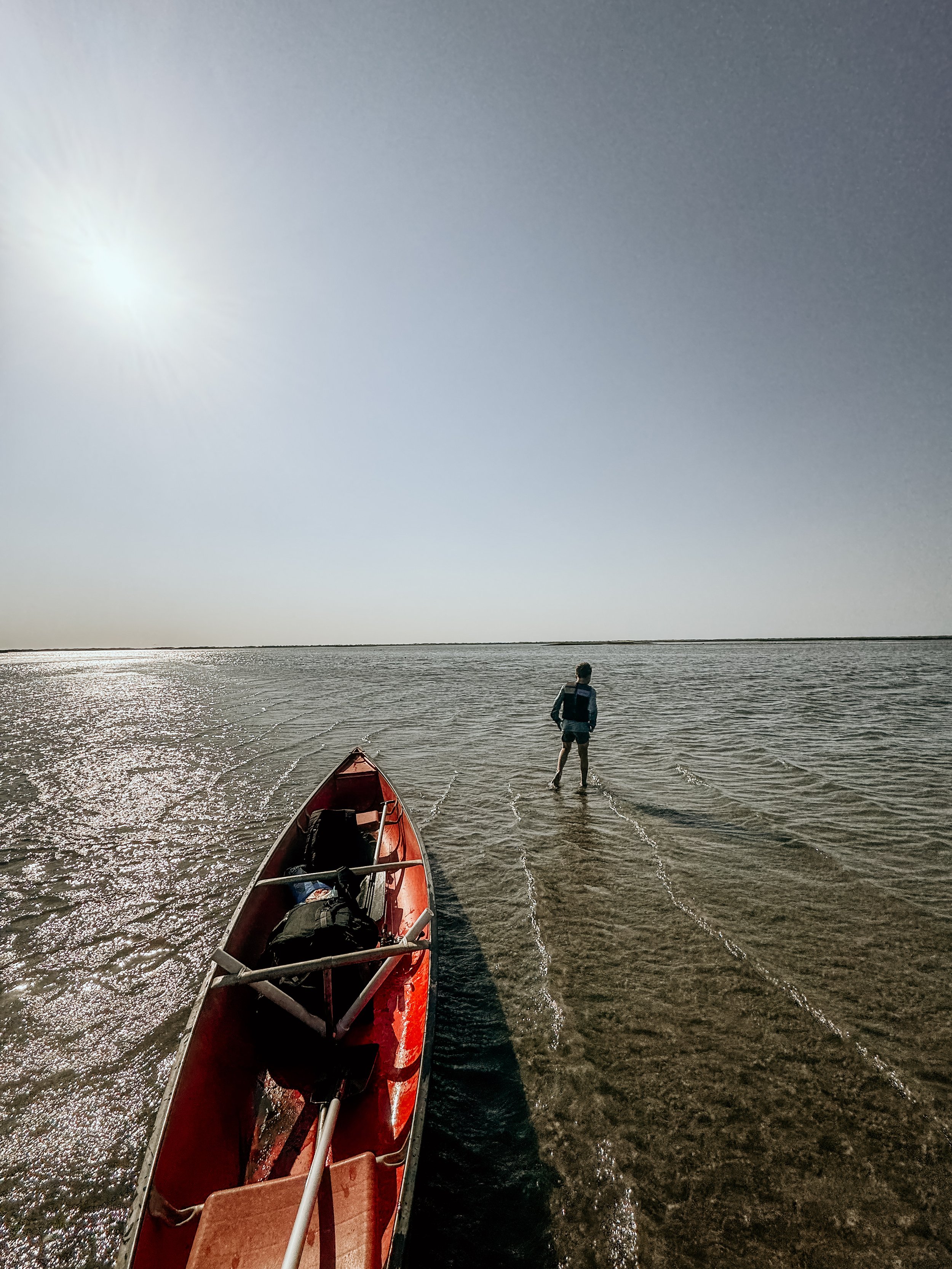 A person standing in shallow water by a red canoe on a calm body of water under a bright sky with the sun shining.