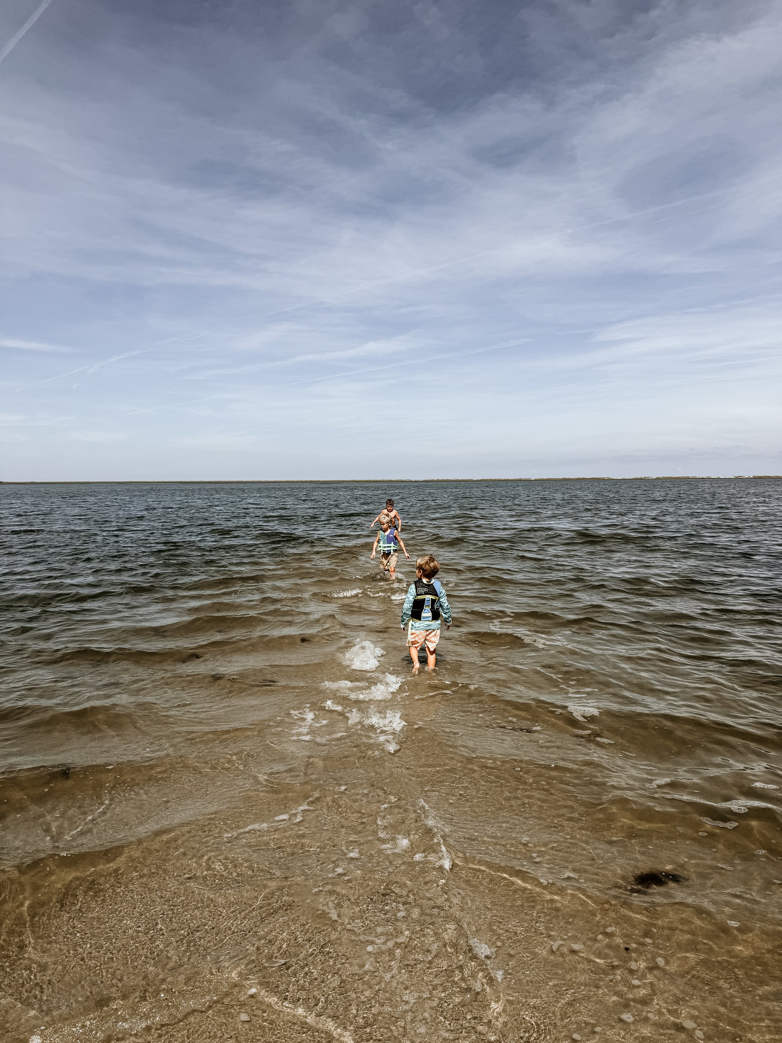 Three children with life jackets walking into the water at a beach on a cloudy day.