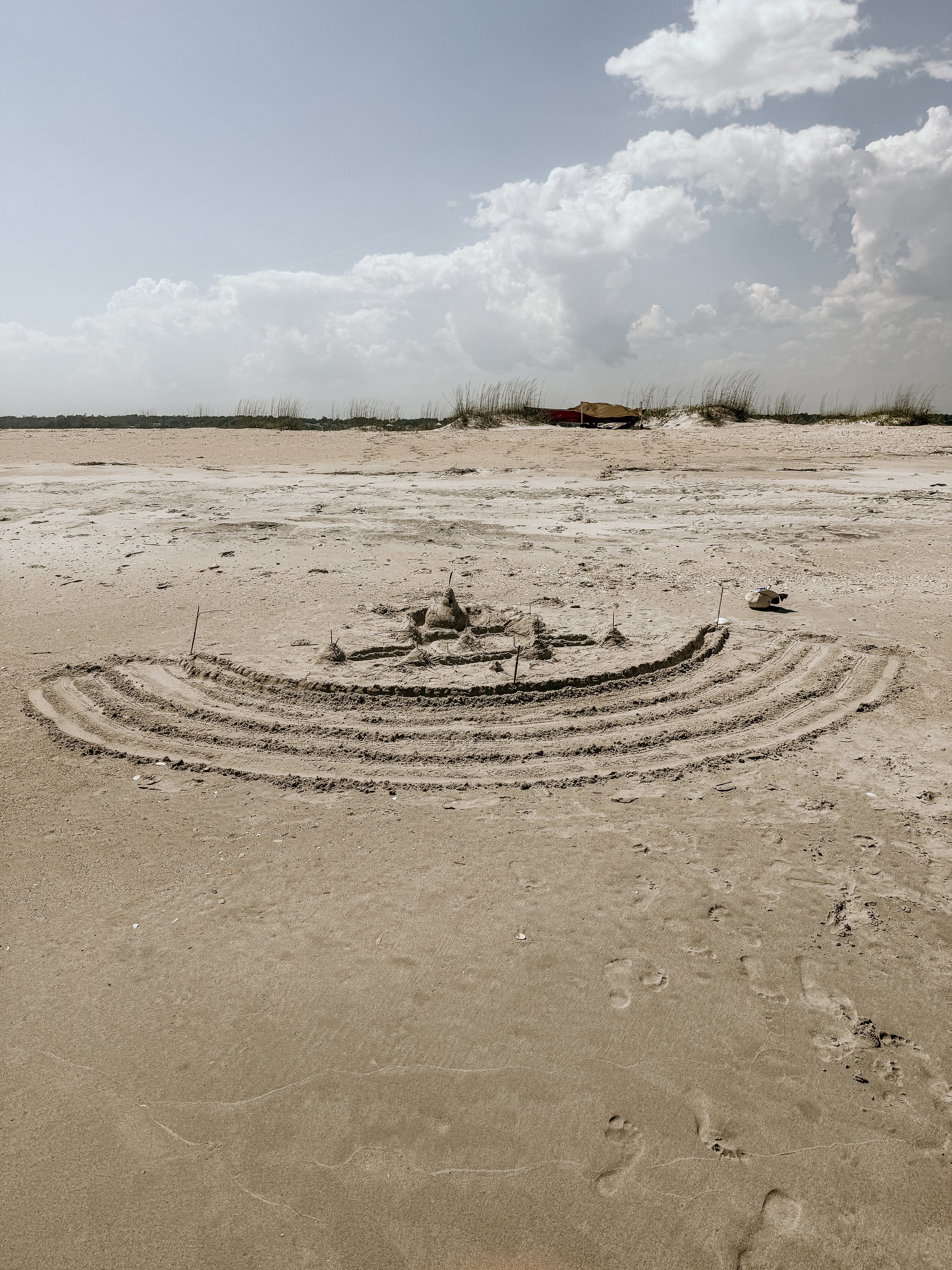 A sandy beach with tire tracks and footprints, dunes with grass, and a cloudy sky.