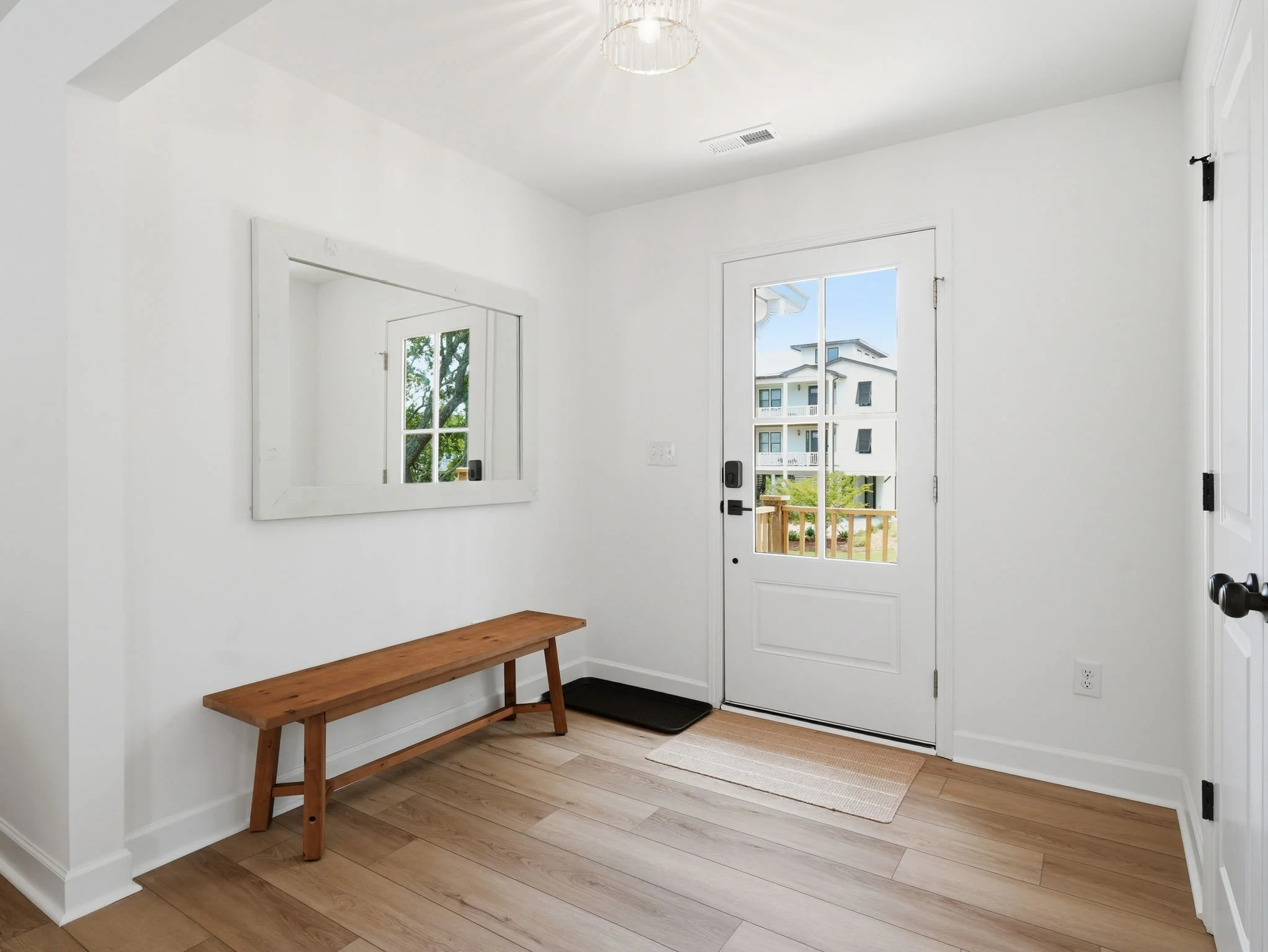 Entryway with white walls, a glass door with a screen, a wooden bench, a rectangular mirror, and a view of a balcony and neighboring buildings outside.