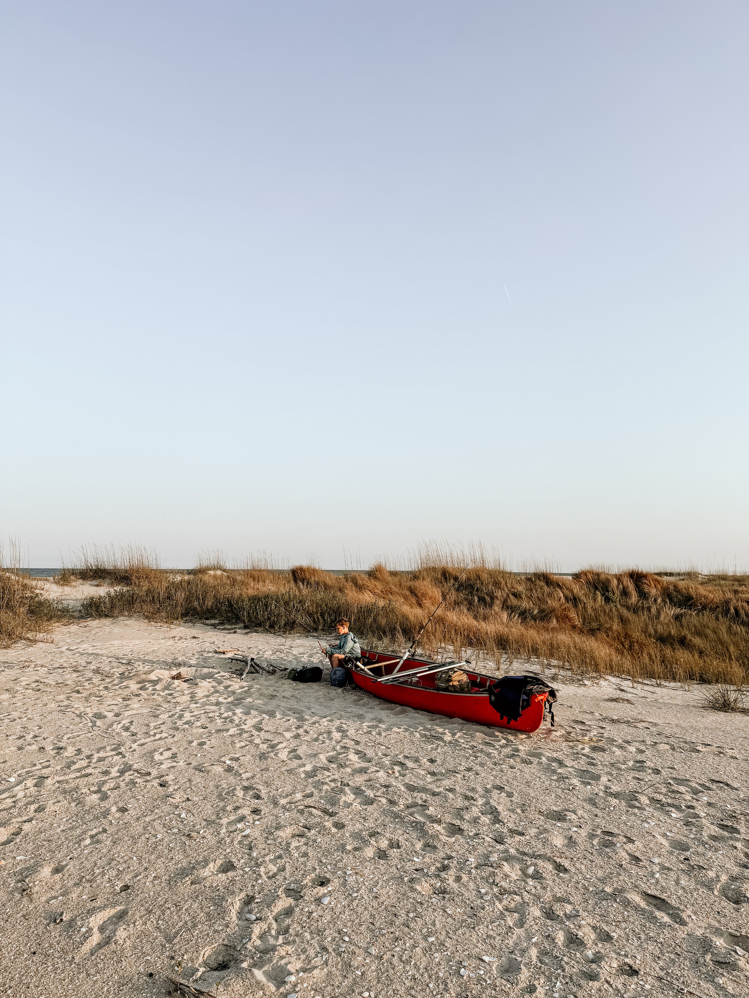 A person sitting near a red kayak on a sandy beach, with grassy dunes and clear blue sky in the background.