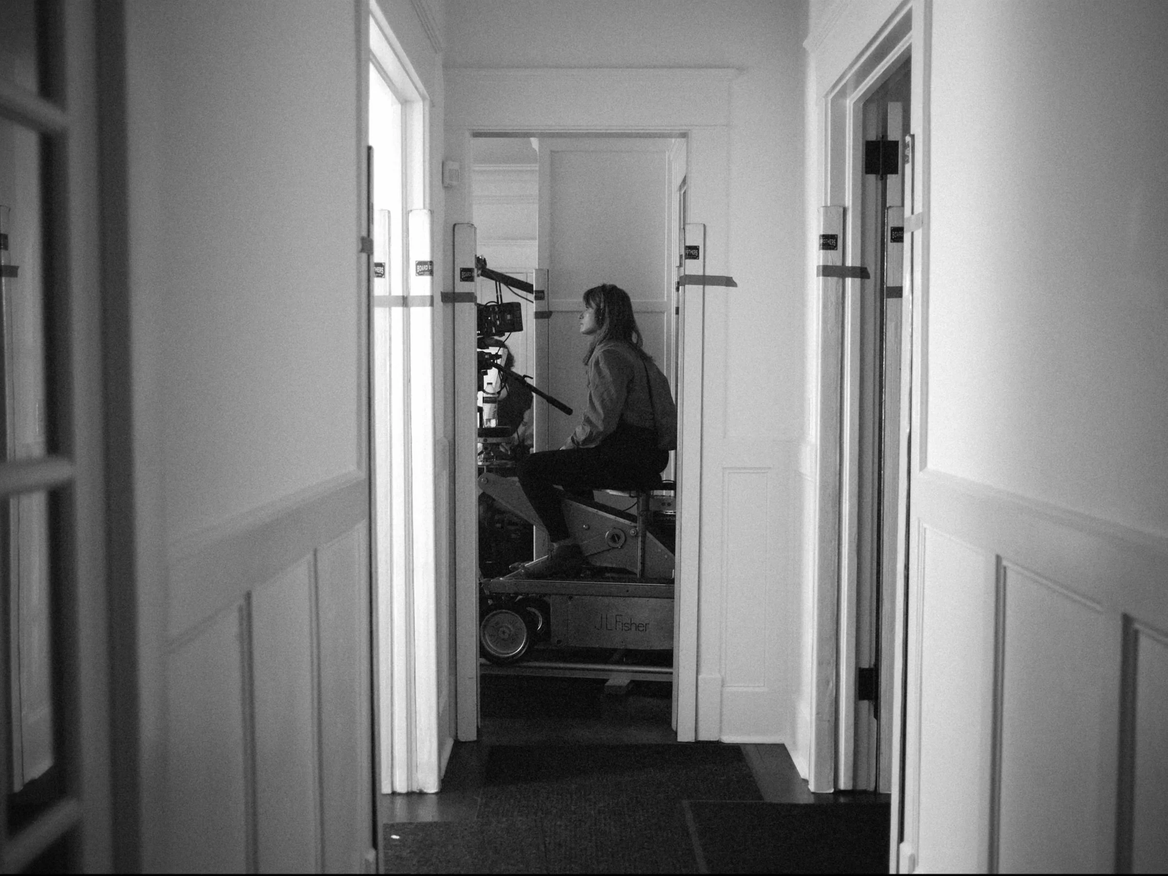 Black-and-white behind-the-scenes image of Lara Aqel operating camera on dolly on film set.