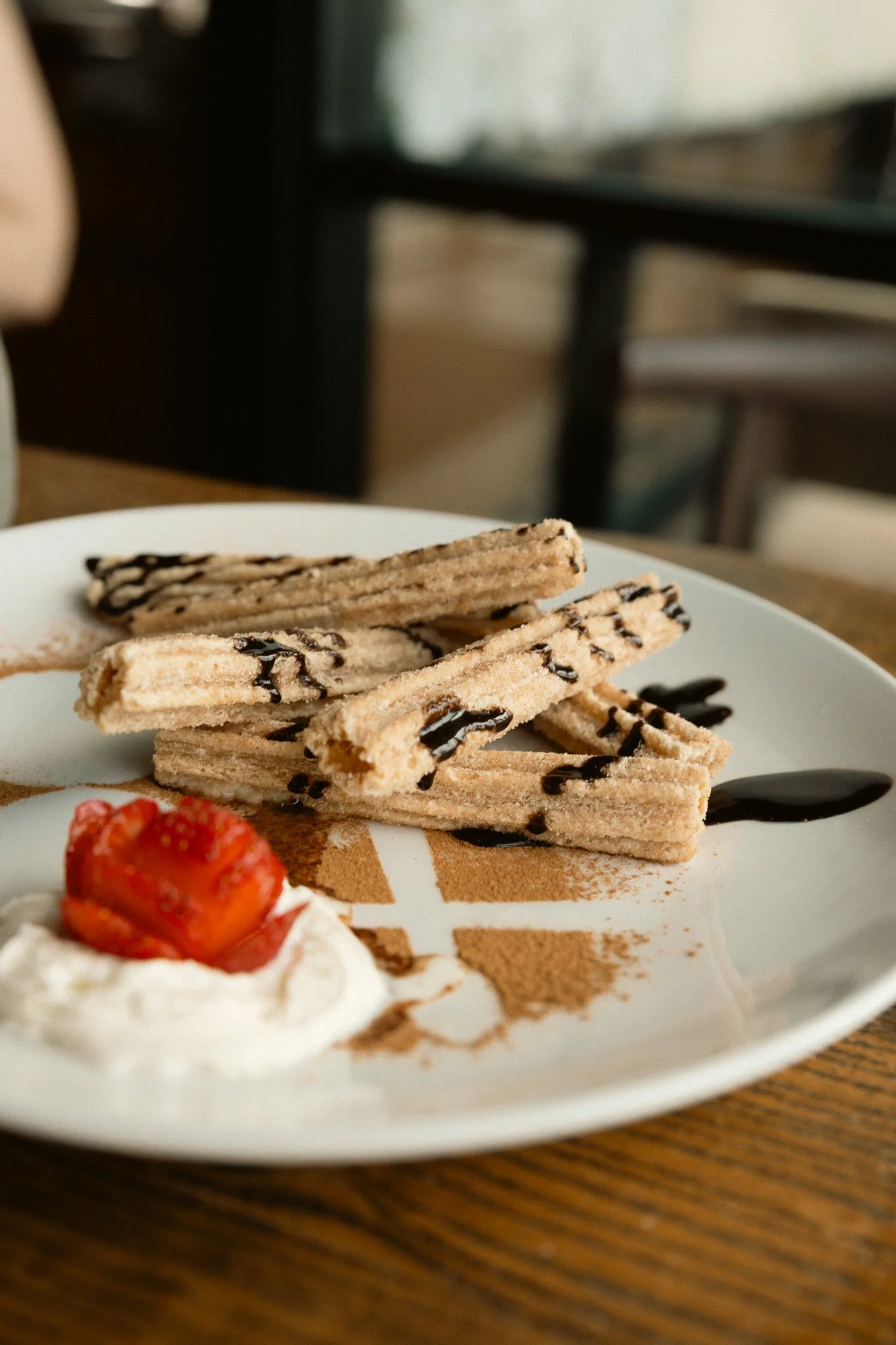Close-up of a dessert plate with cookies drizzled with chocolate, a dollop of whipped cream topped with a strawberry, and cocoa powder on the plate. The plate is on a wooden table, with a blurred background.