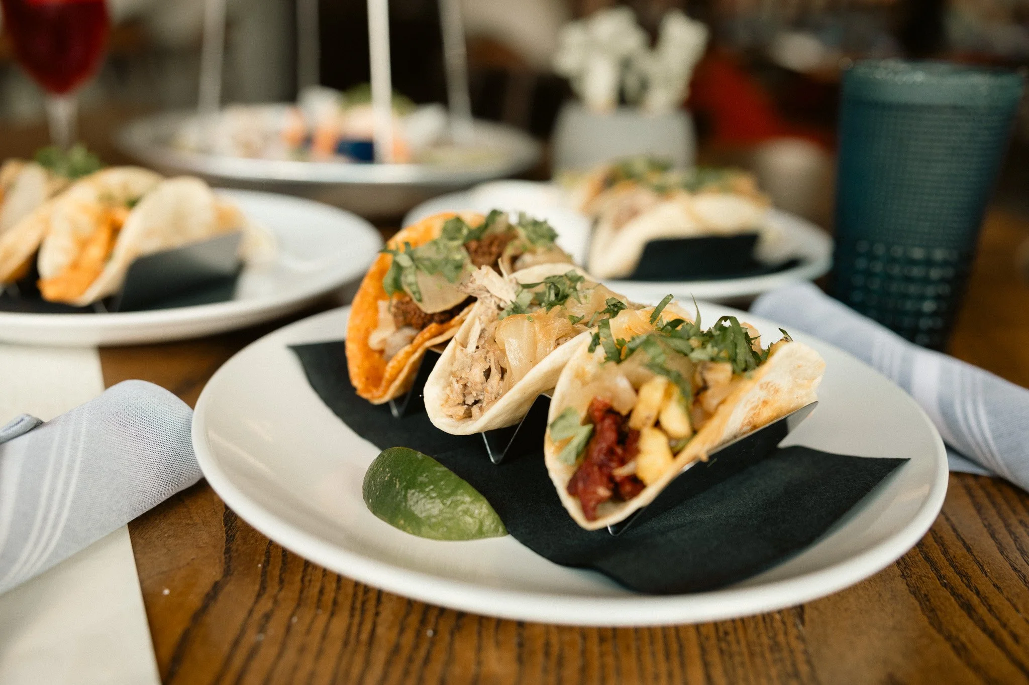 Close-up of three tacos filled with meat, vegetables, and herbs, served on a white plate with a lime wedge, on a wooden table with white napkins nearby.