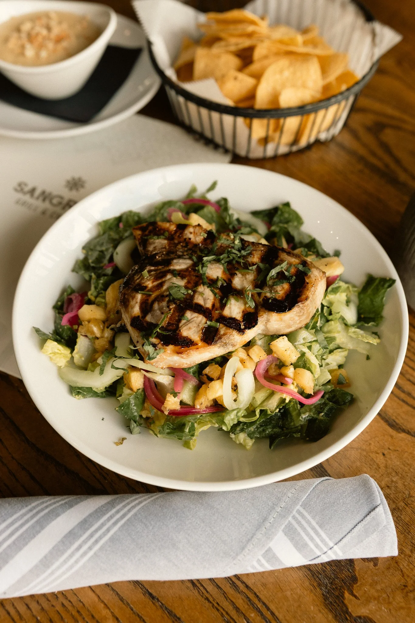 A white bowl of mixed salad topped with grilled chicken and drizzled with balsamic glaze, with a basket of potato chips in the background.
