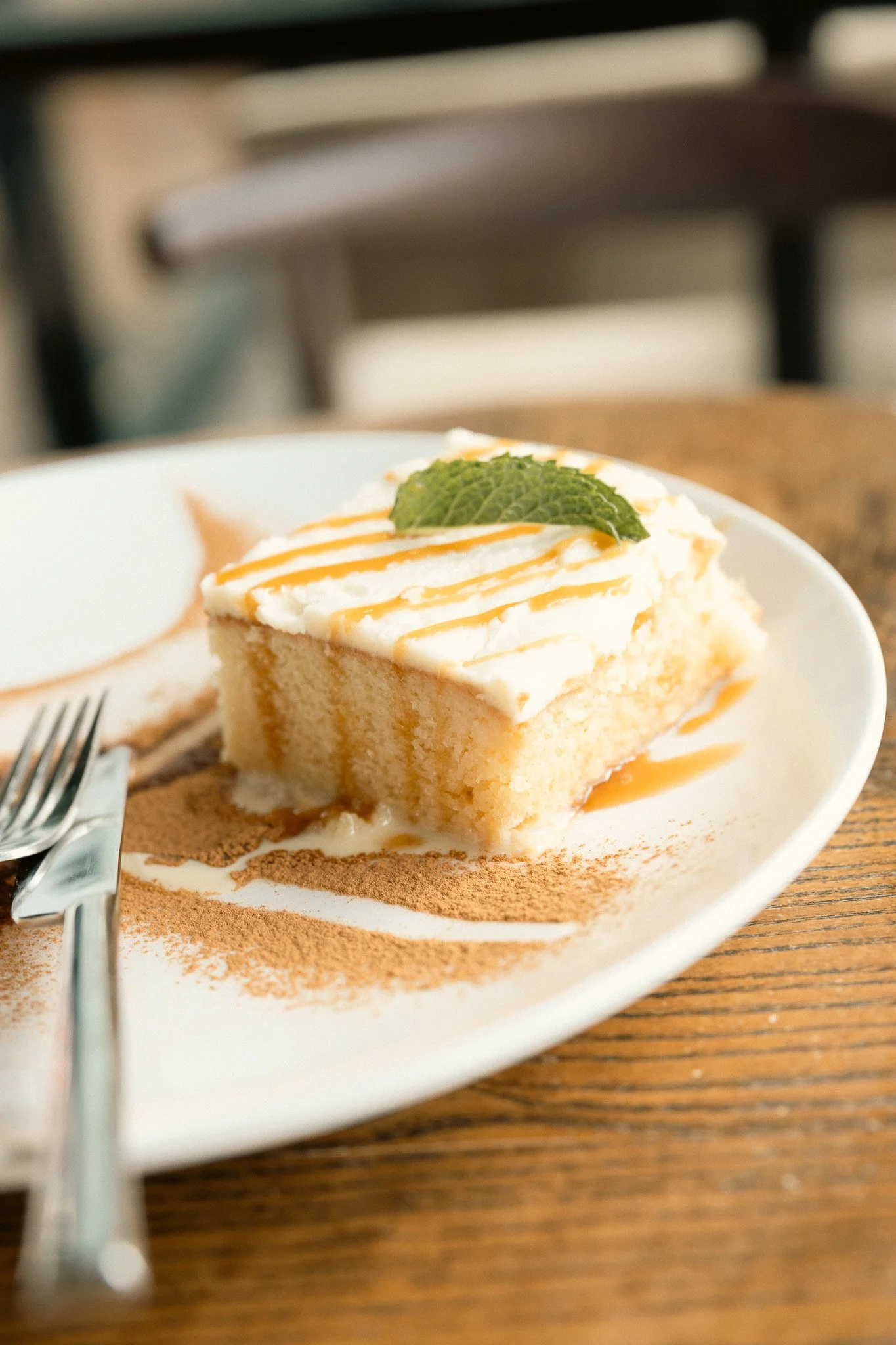 A slice of vanilla cake with white frosting, topped with a mint leaf and orange caramel drizzle, served on a white plate with cocoa powder and cinnamon sprinkled on it, placed on a wooden table.